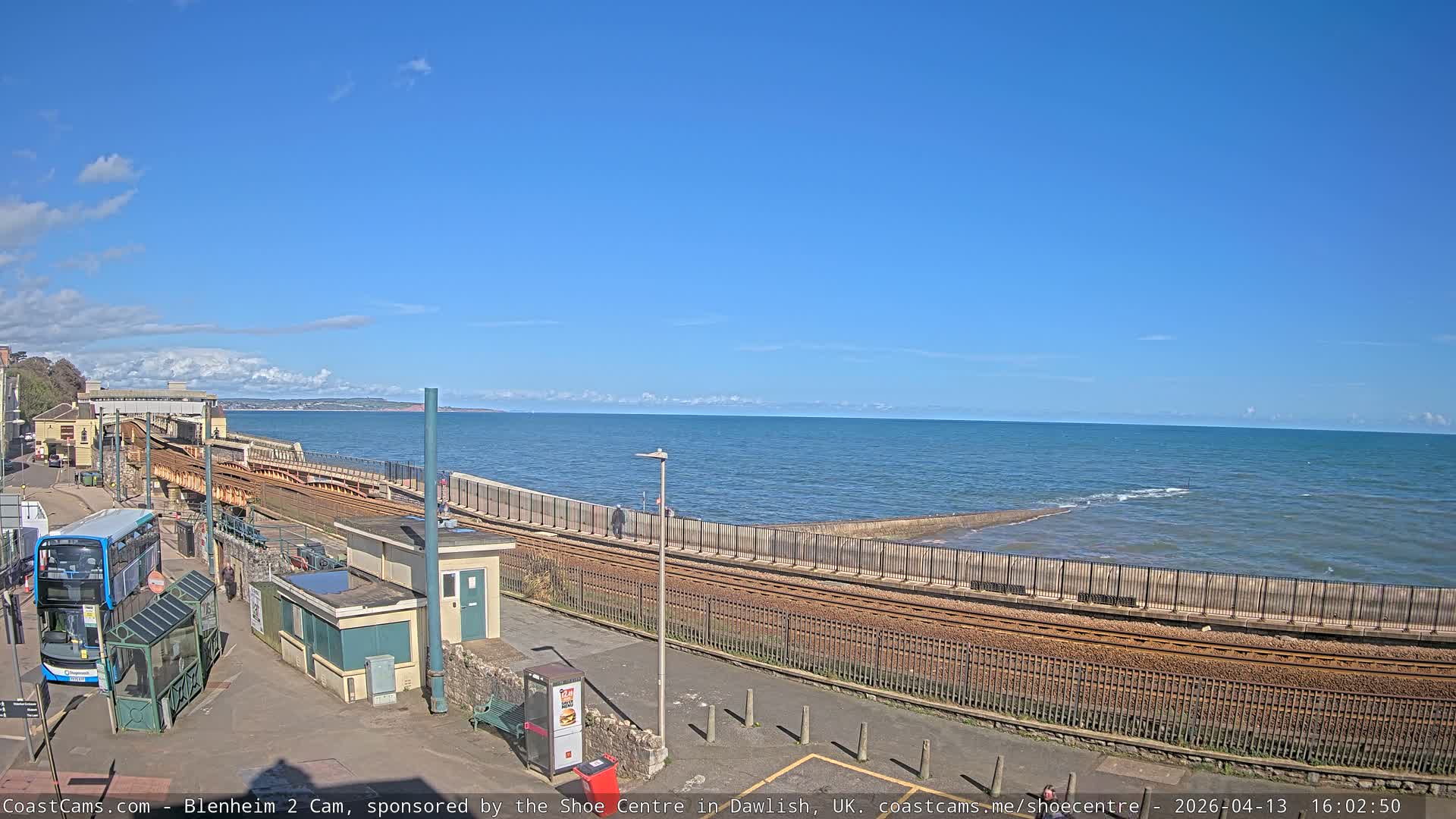 On a bright, clear morning, a coastal scene reveals train tracks and a concrete sea wall bordering a calm ocean with small waves, while a person in an orange jacket scooters along a paved path next to a town built on a hillside.