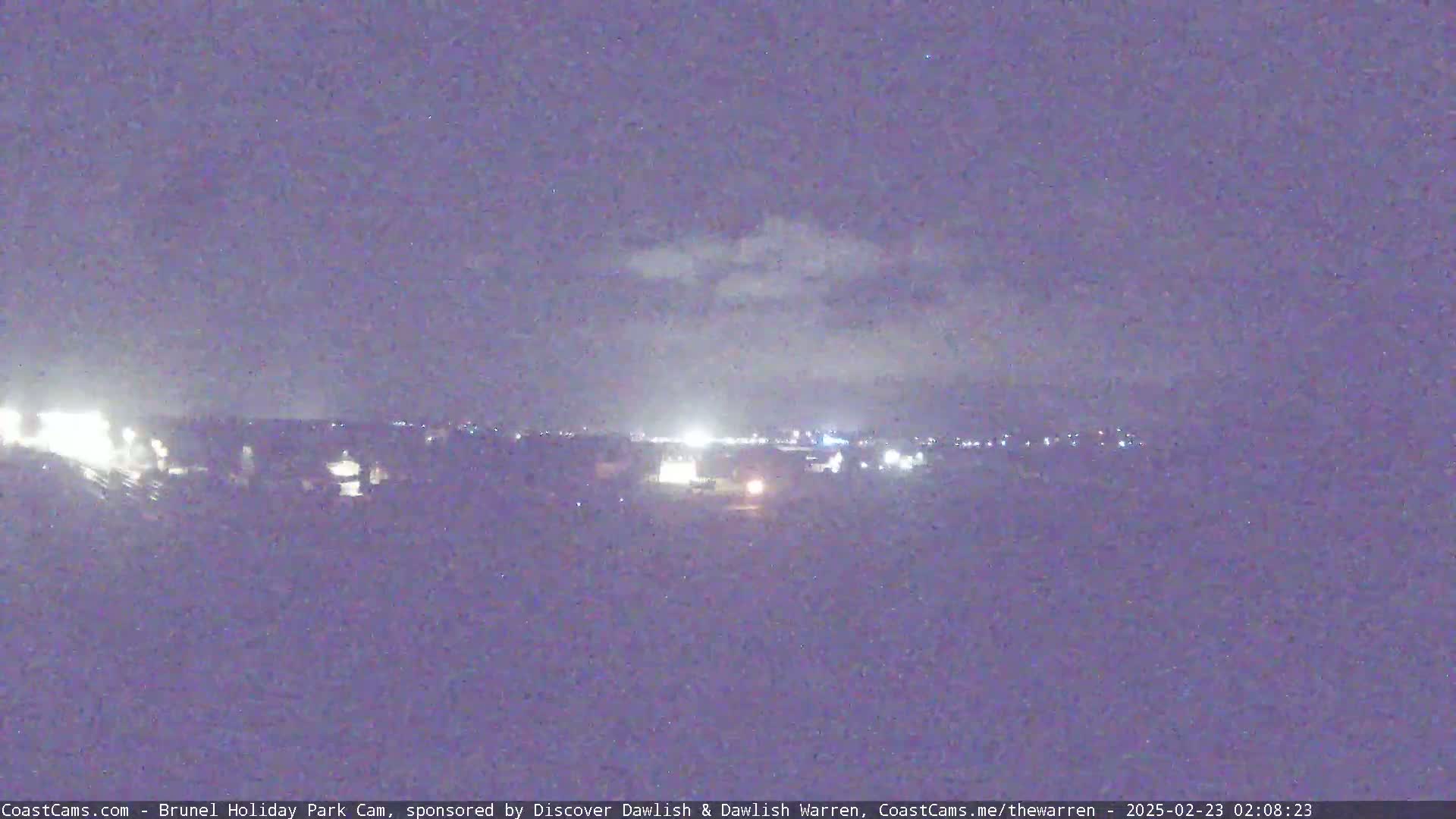 A nighttime, dimly lit landscape with a few lights visible in the distance under a cloudy sky.