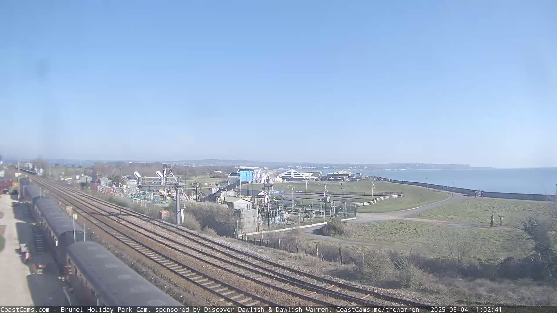A seaside amusement park sits beside a railway line and a calm ocean under a clear blue sky.