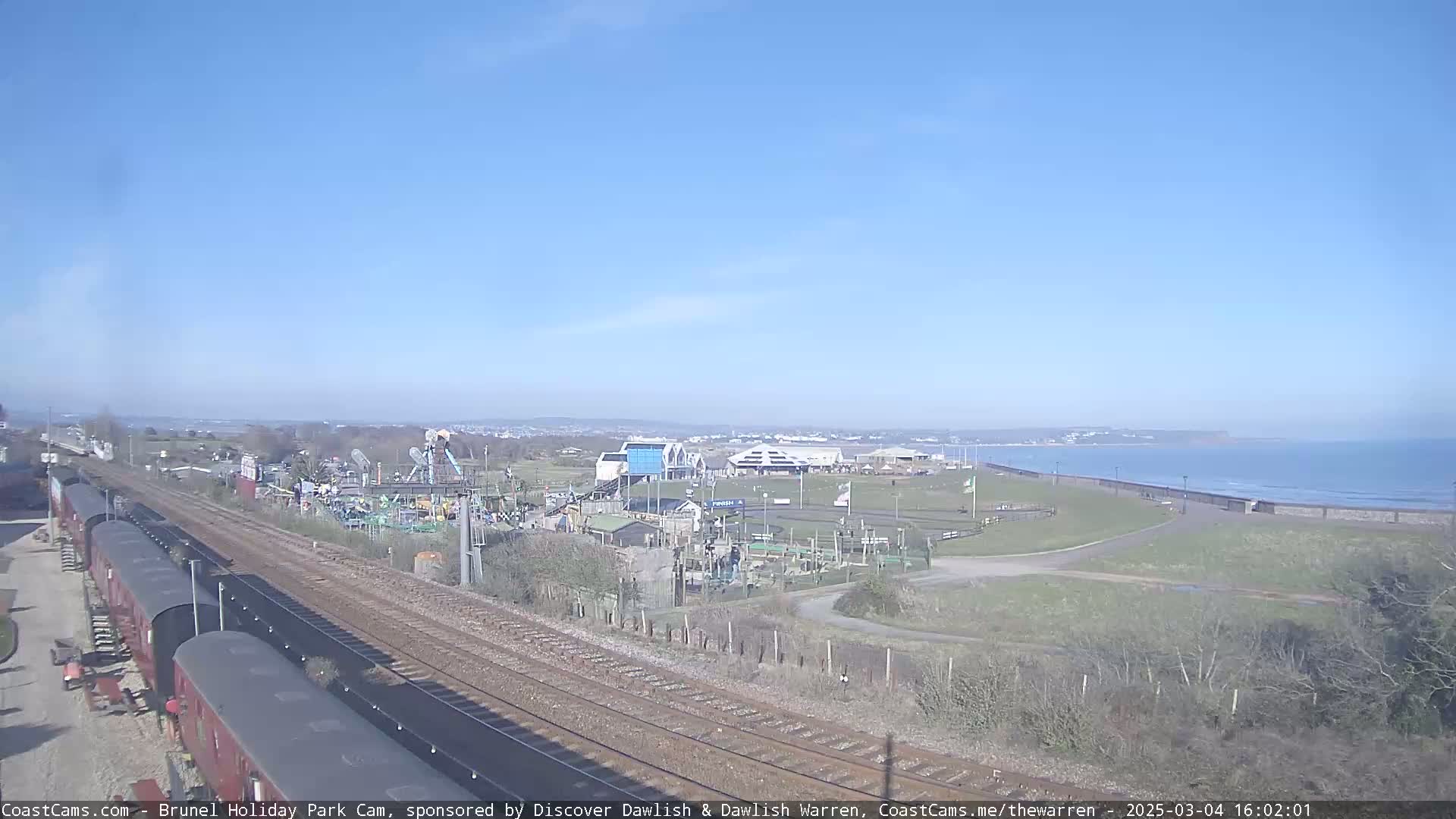 A sunny day reveals a seaside amusement park next to a train track and a calm ocean.