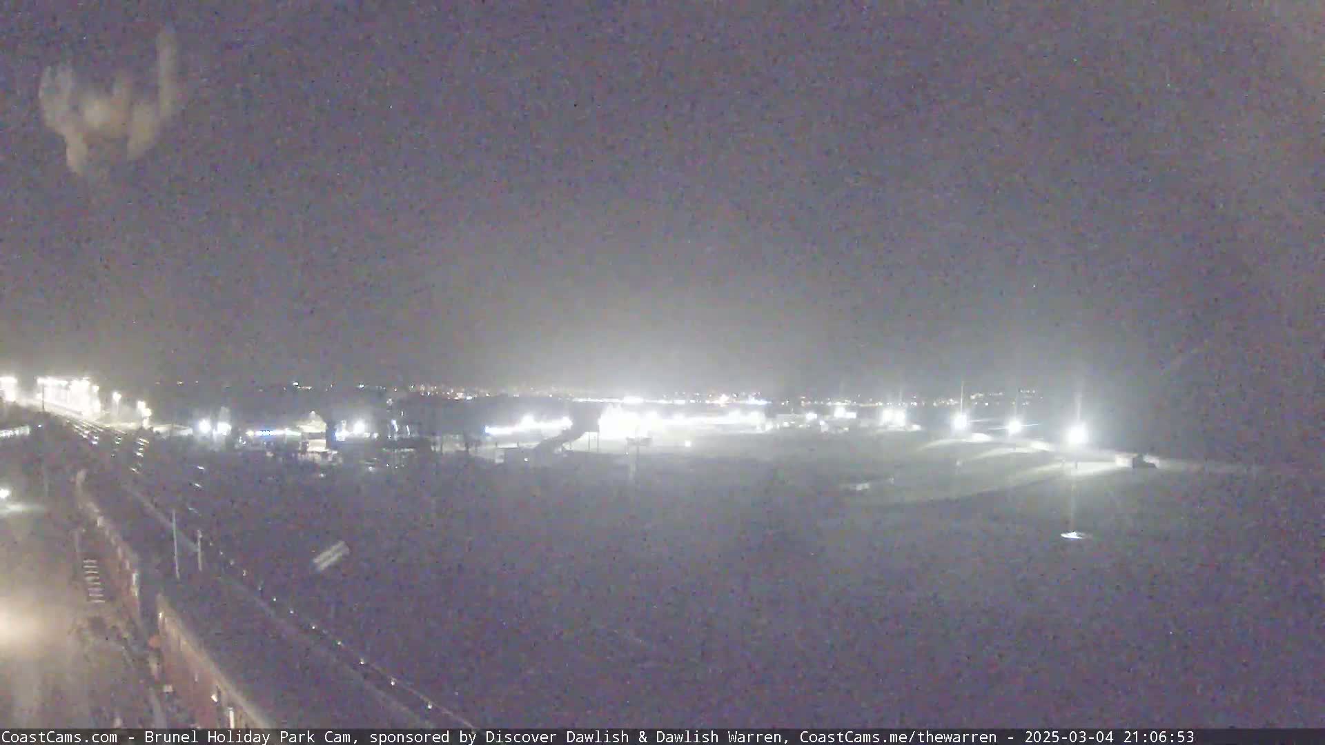 A nighttime, low-light view of a large, flat, mostly empty area with some distant lights and a roadway visible in the foreground.