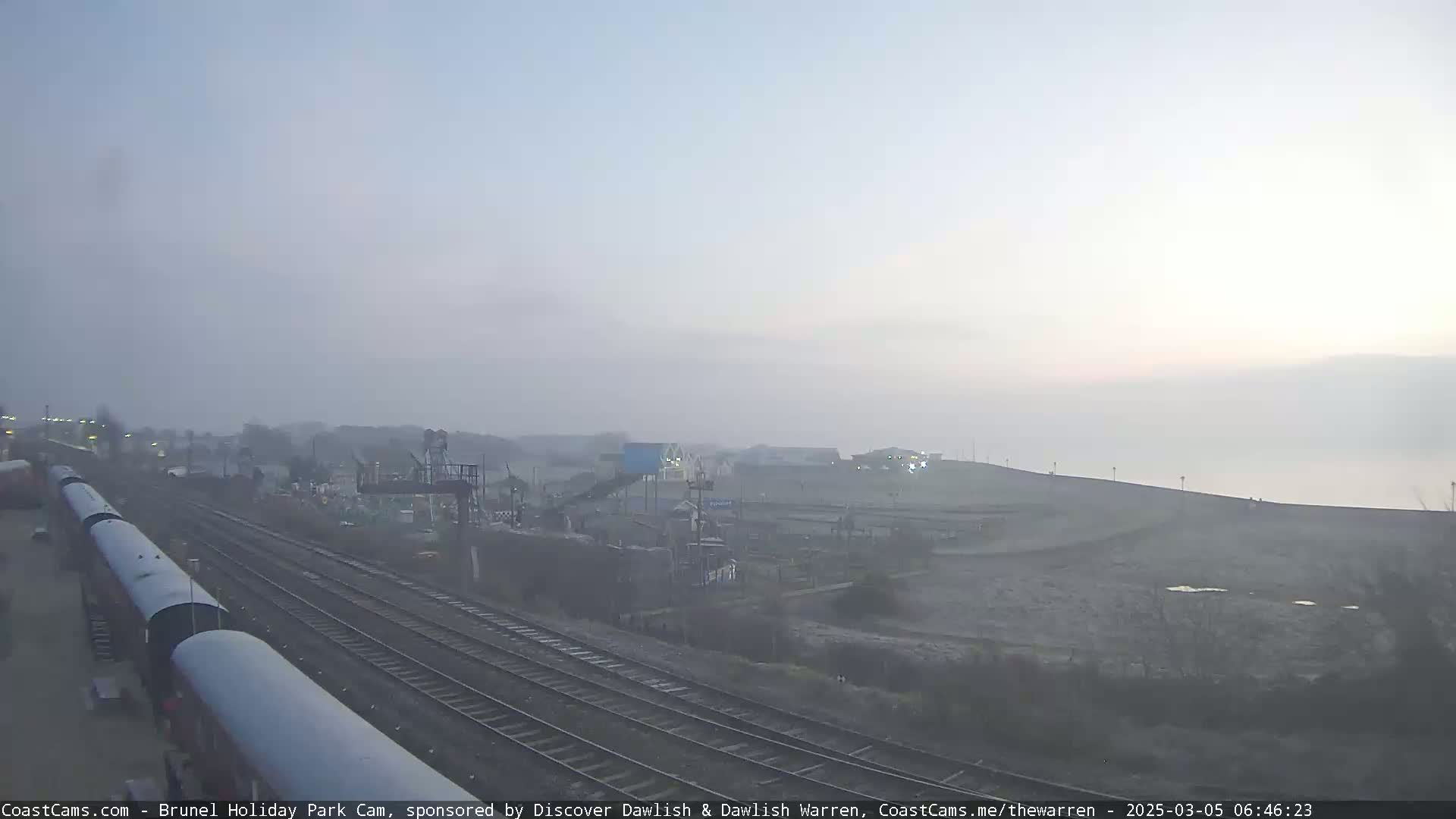 A foggy sunrise scene shows a train and railway tracks next to a seaside amusement park and a sloping expanse of land leading to the water.