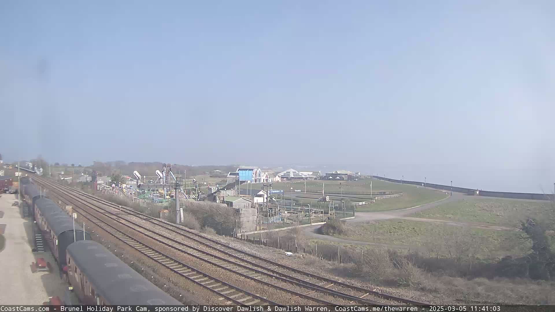 A long, elevated view shows a railway track next to a playground and other recreational facilities, all situated near a coastline on a clear, sunny day.