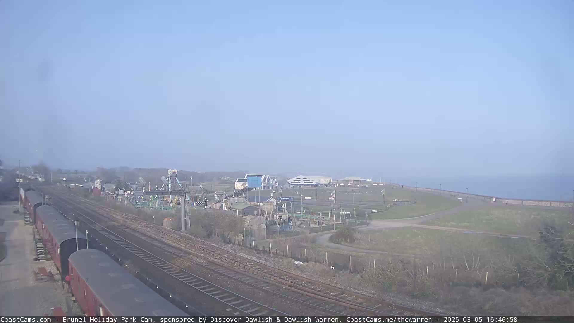 A clear day reveals a seaside amusement park near train tracks, with a calm body of water in the background.