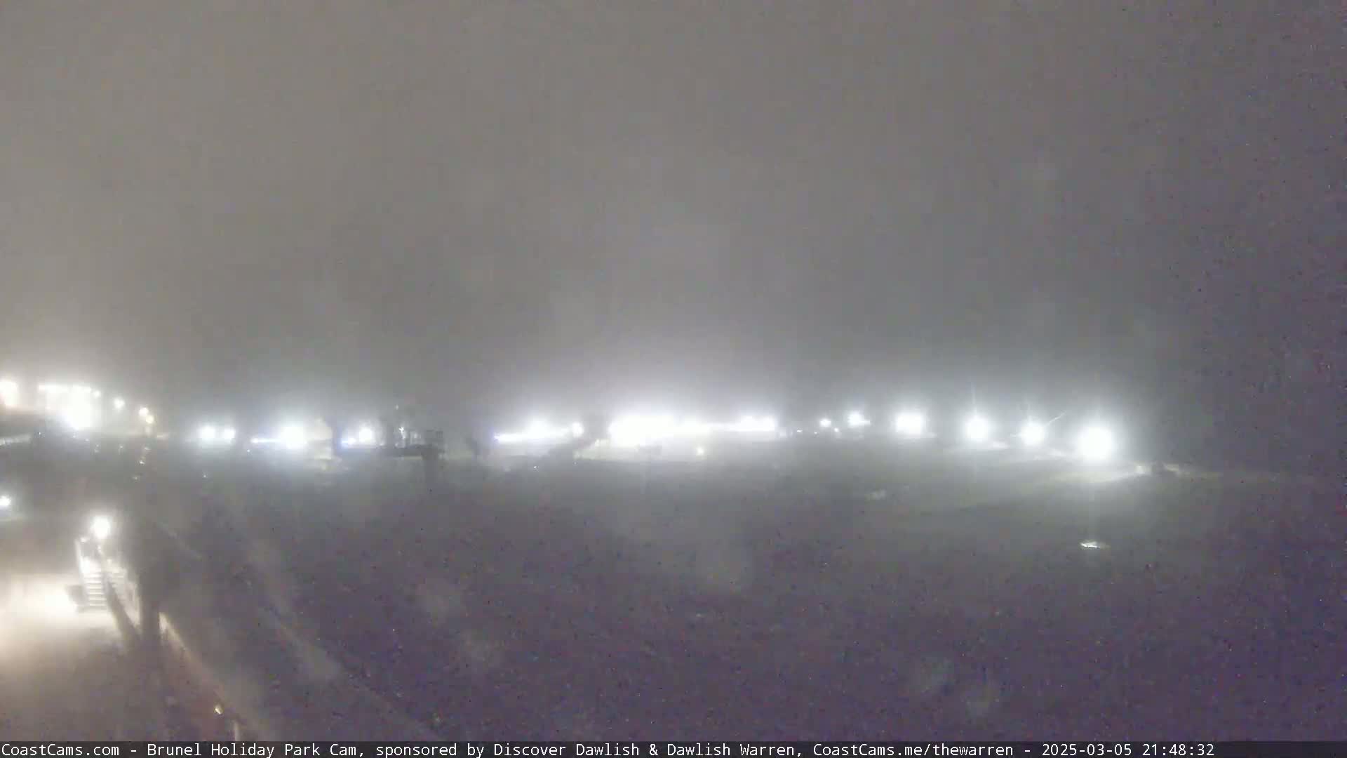 A foggy night scene shows a dimly lit area with several distant lights, possibly from buildings or vehicles.