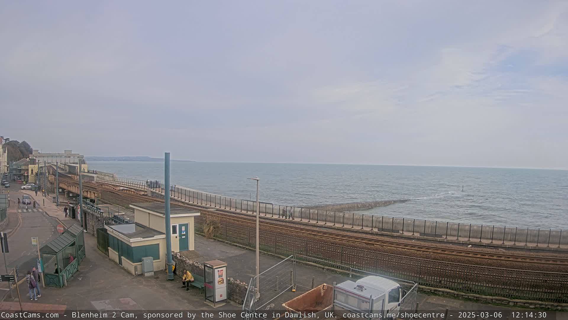 A coastal scene shows a train track running alongside a sea wall, with a calm sea extending to the horizon under a cloudy sky.