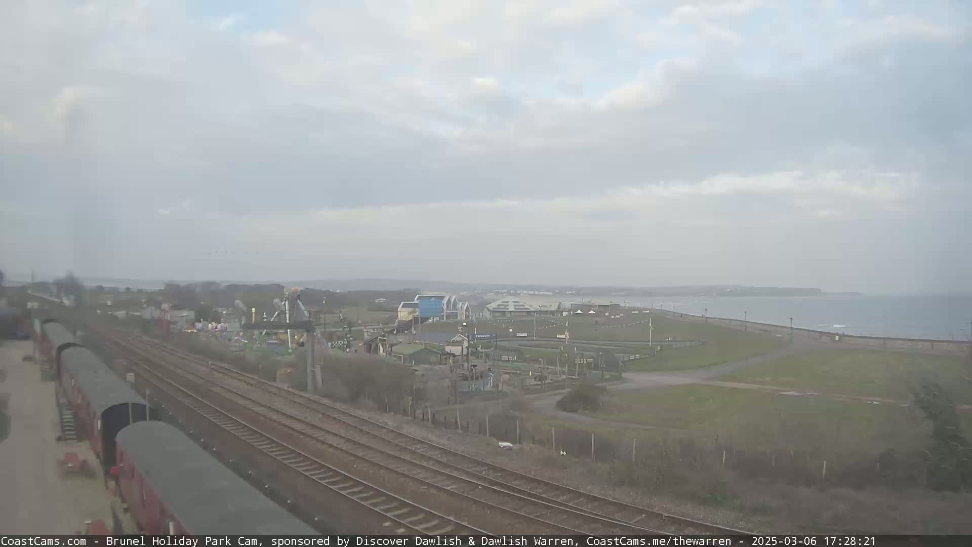 A seaside amusement park is visible beyond railroad tracks and freight cars under an overcast sky.