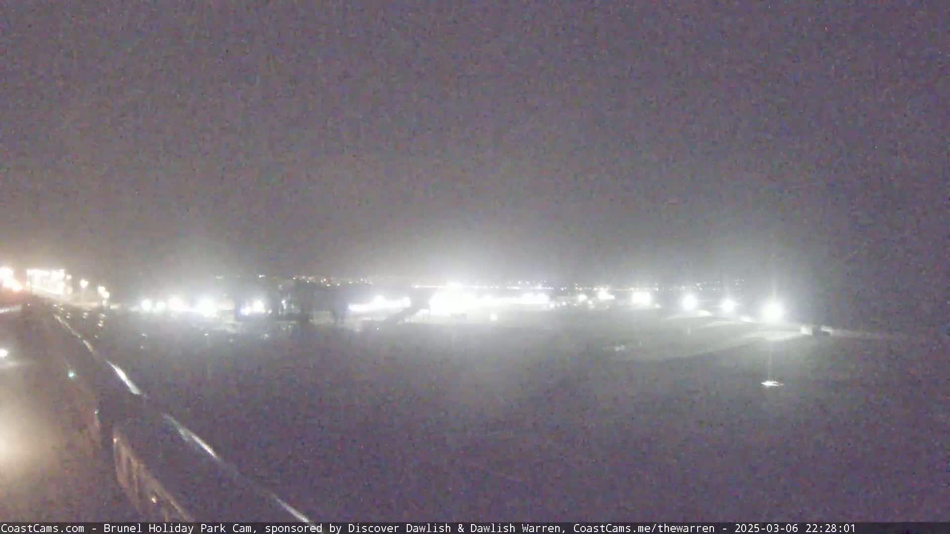 A nighttime, hazy view of a brightly lit area, possibly a parking lot or outdoor facility, with a dark road in the foreground.