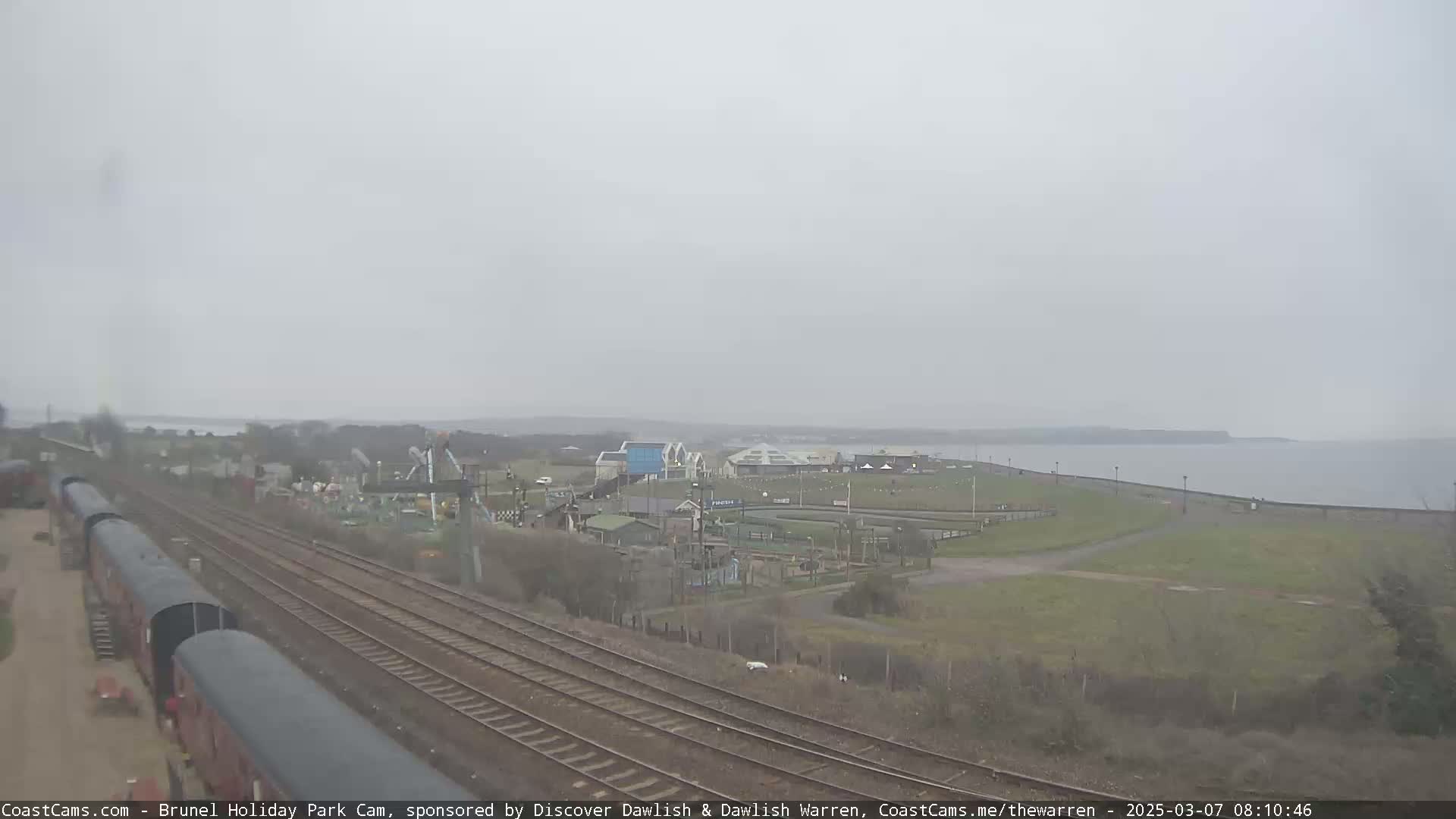 A cloudy day reveals train tracks leading to a seaside holiday park with a body of water in the background.