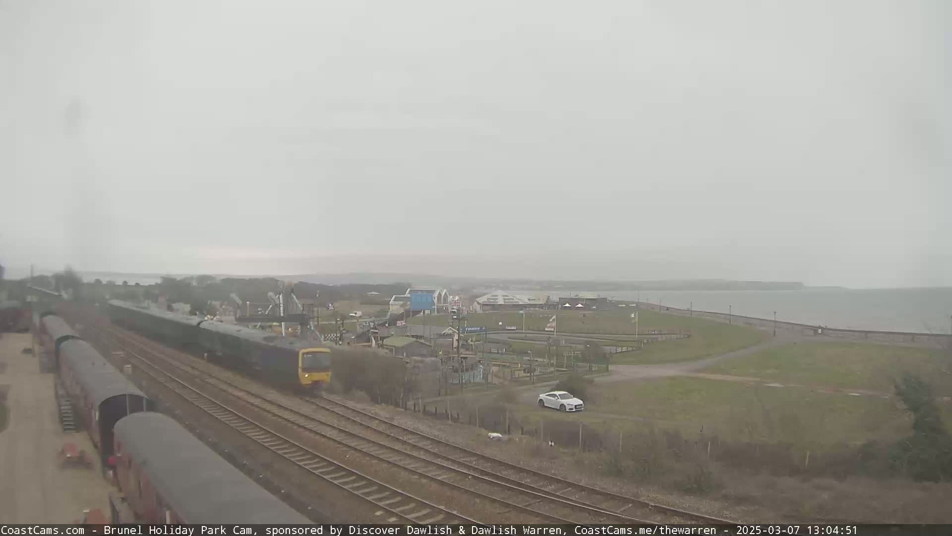 A green passenger train travels along a railway line near a coastline under an overcast sky, with a seaside park and a car visible in the distance.