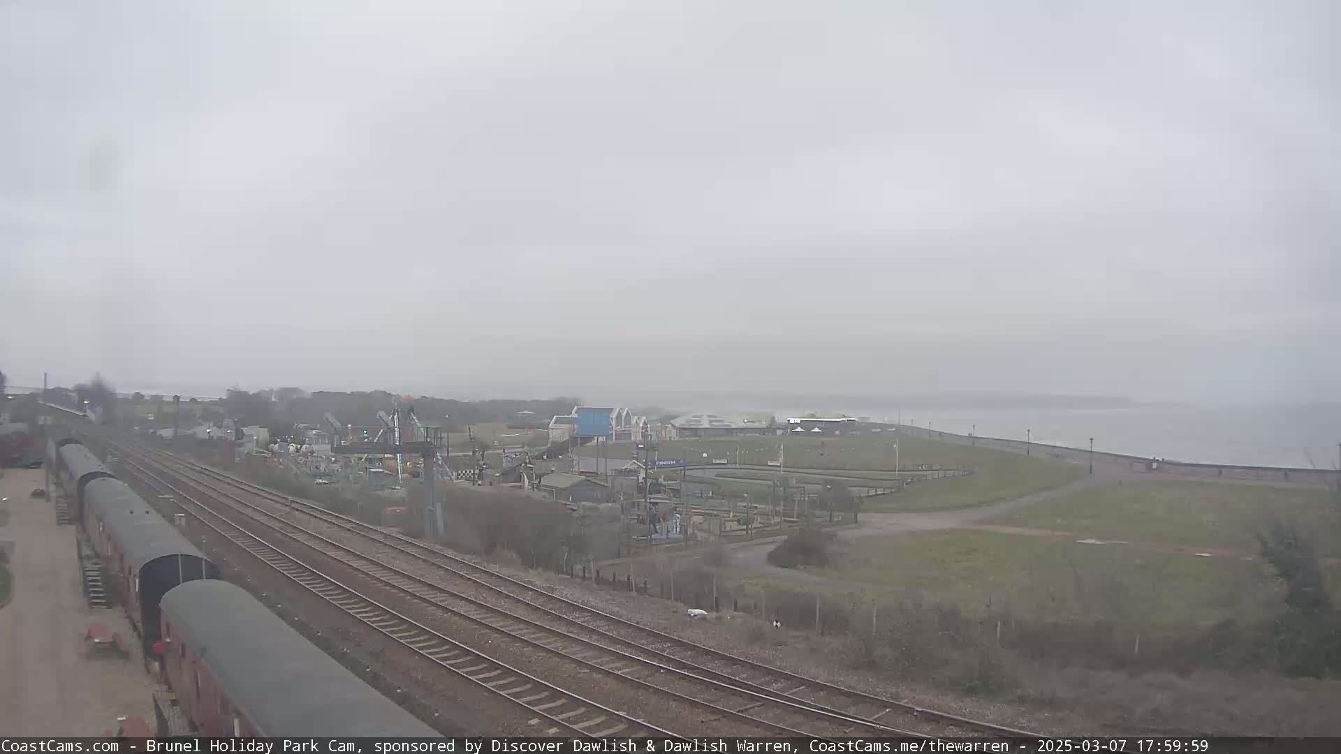 A foggy view of a coastal area shows train tracks running alongside a holiday park with amusement park rides near a body of water.