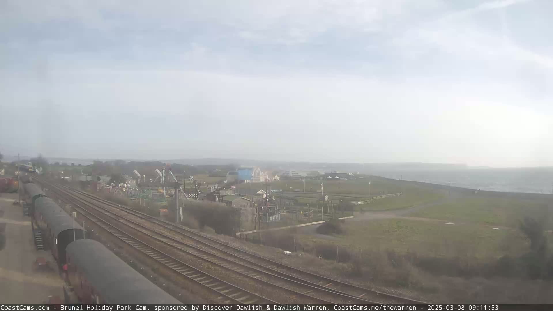 A hazy, overcast day reveals railway tracks leading to a coastal area with a holiday park, featuring a playground and a body of water in the distance.
