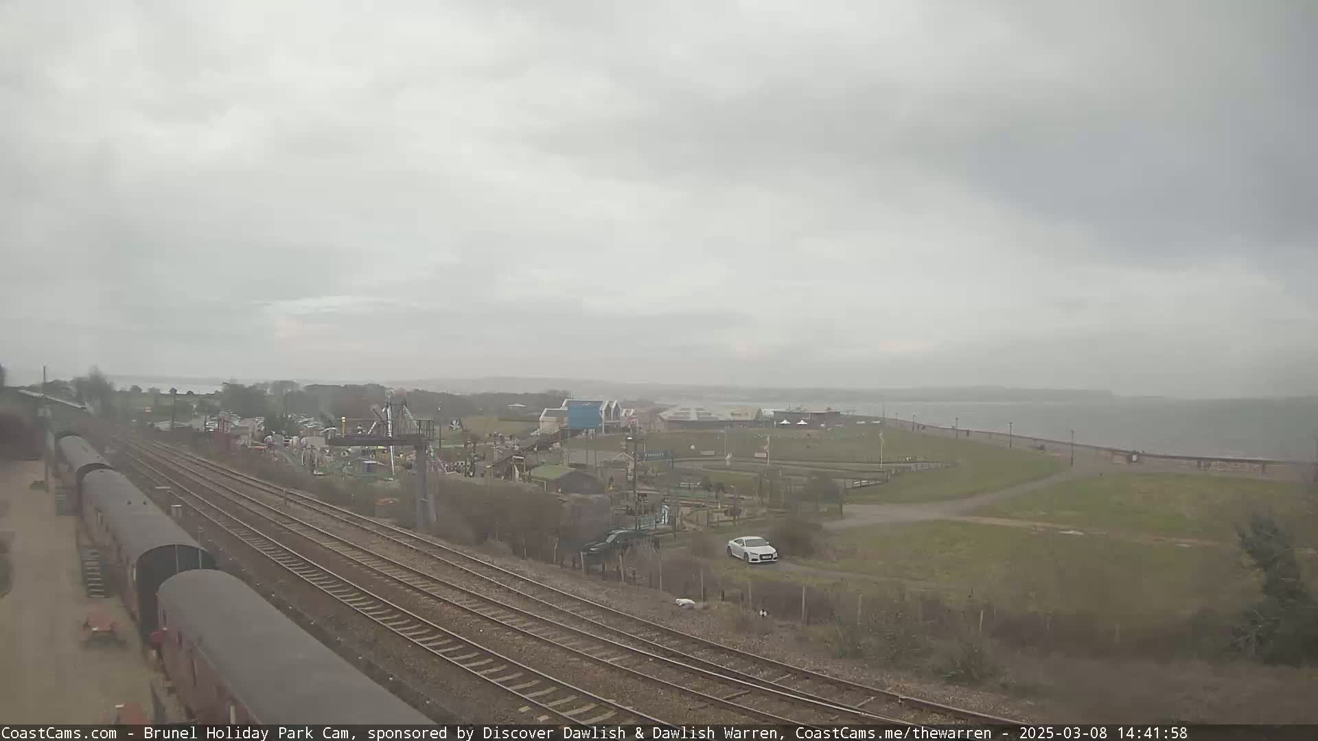 A mostly cloudy, overcast day shows a view of a train track next to a holiday park near a body of water.