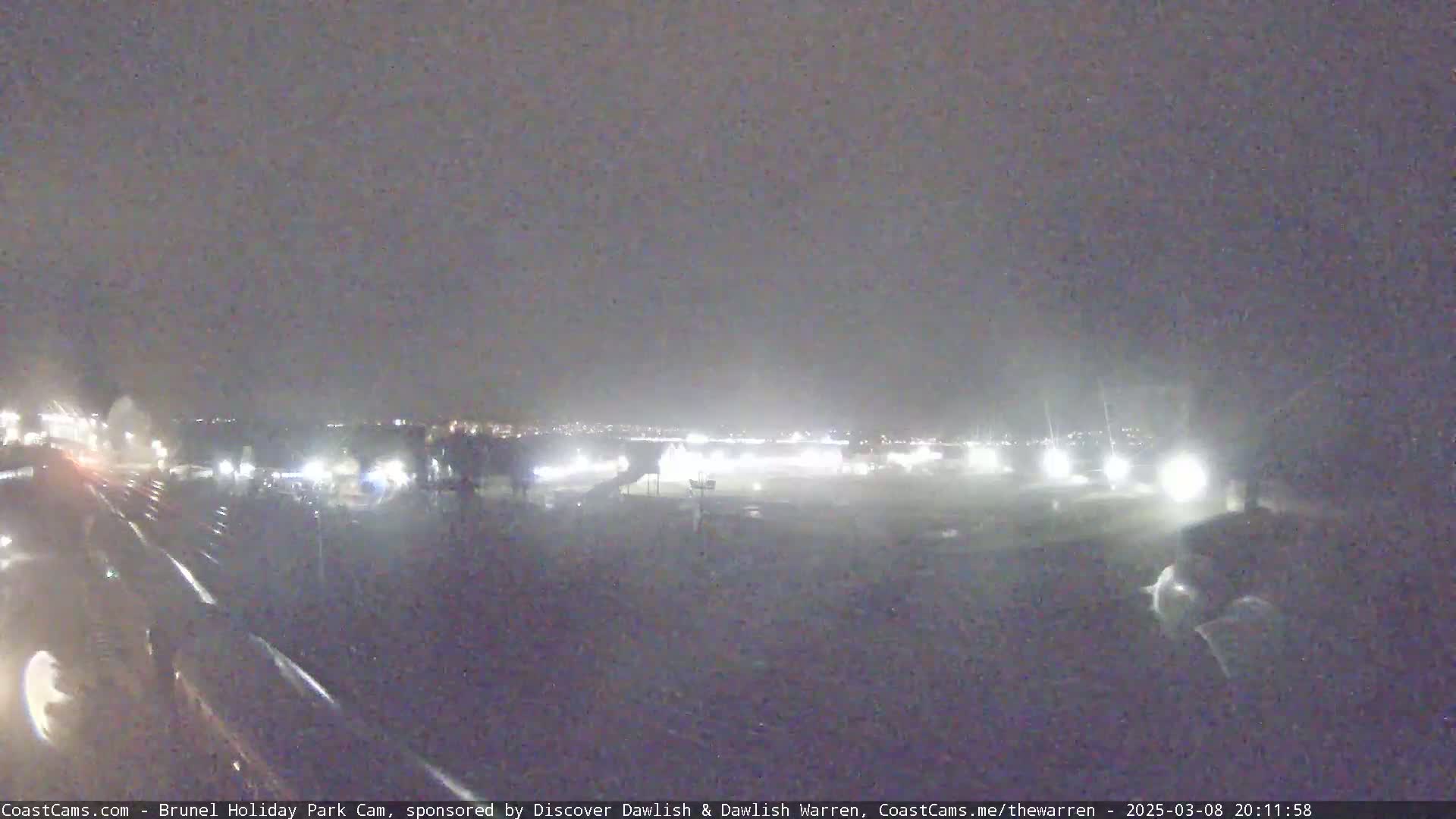 A nighttime, rainy view of a dark, empty parking lot with distant lights and a few indistinct figures.