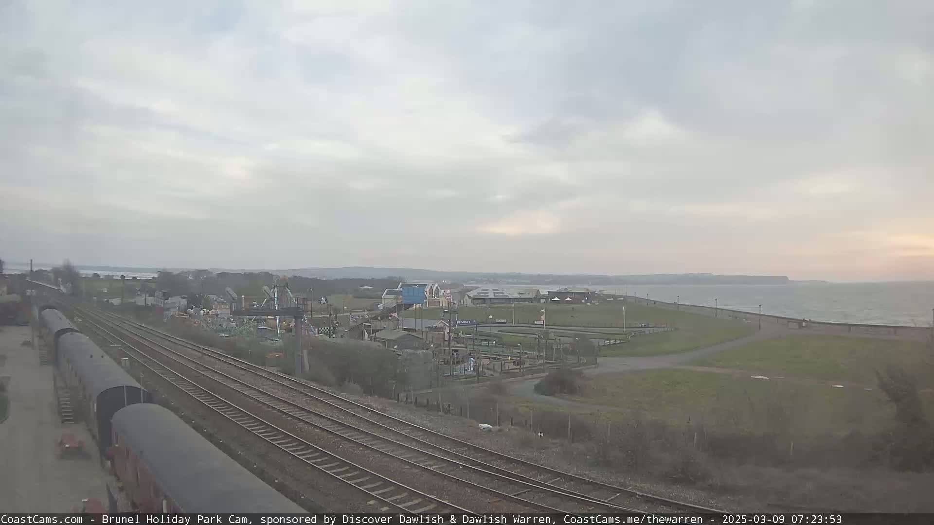 A coastal amusement park is visible beyond railway tracks, under an overcast sky with a calm sea in the background.