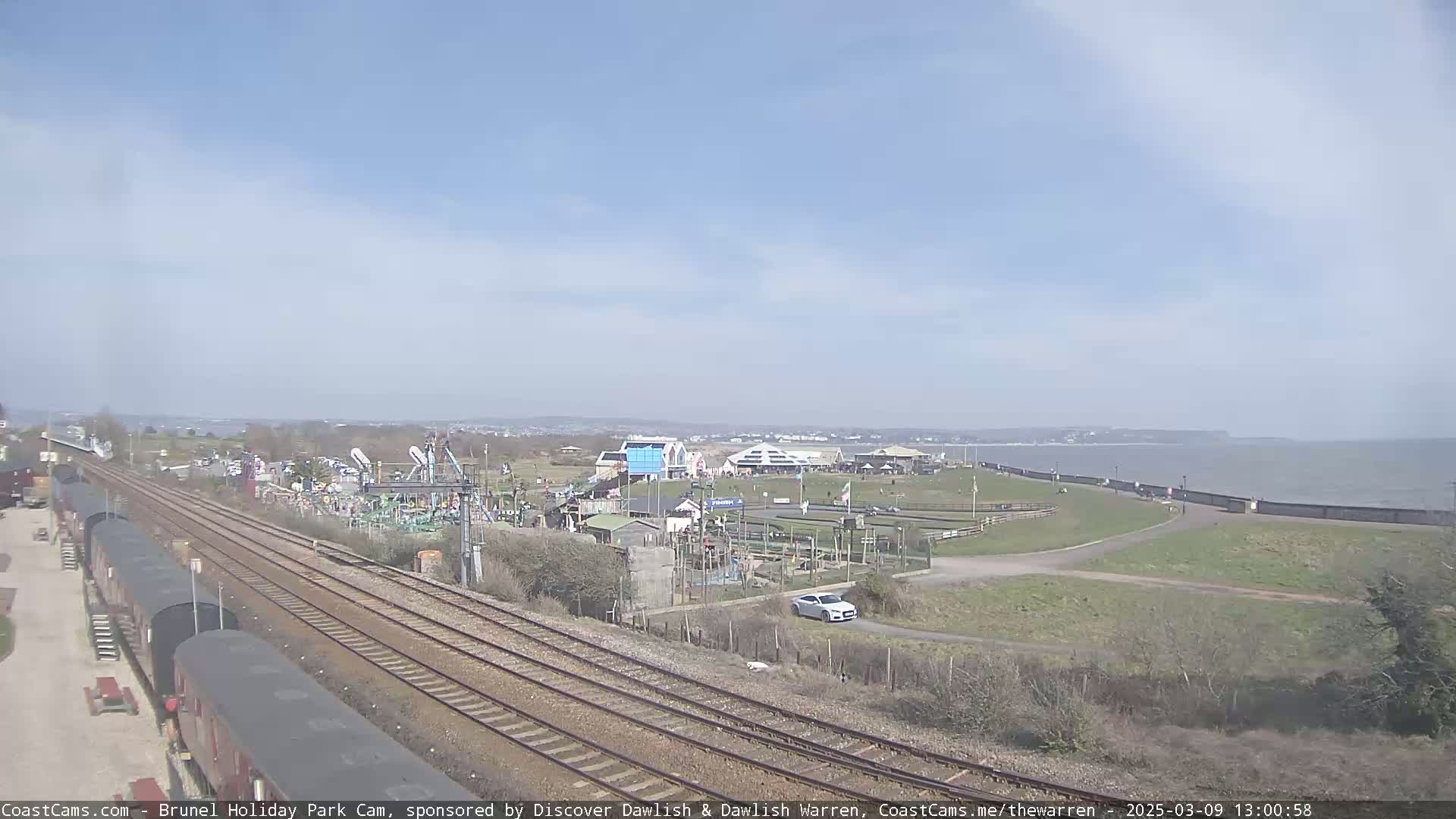 A seaside amusement park is visible beyond railway tracks and several dark-colored train cars under a clear, sunny sky.