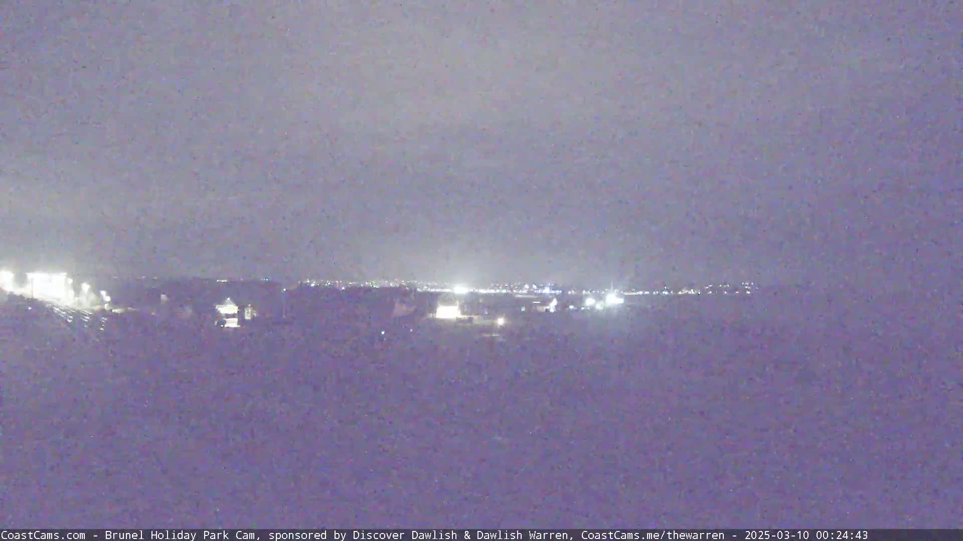 A nighttime long shot shows a distant cityscape illuminated by lights under an overcast sky.