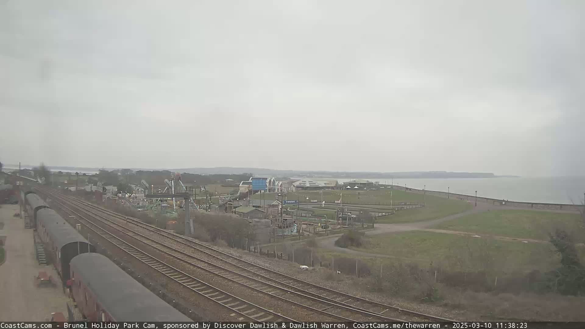 Under an overcast sky, a view shows railroad tracks leading toward a seaside amusement park with a body of water visible in the distance.