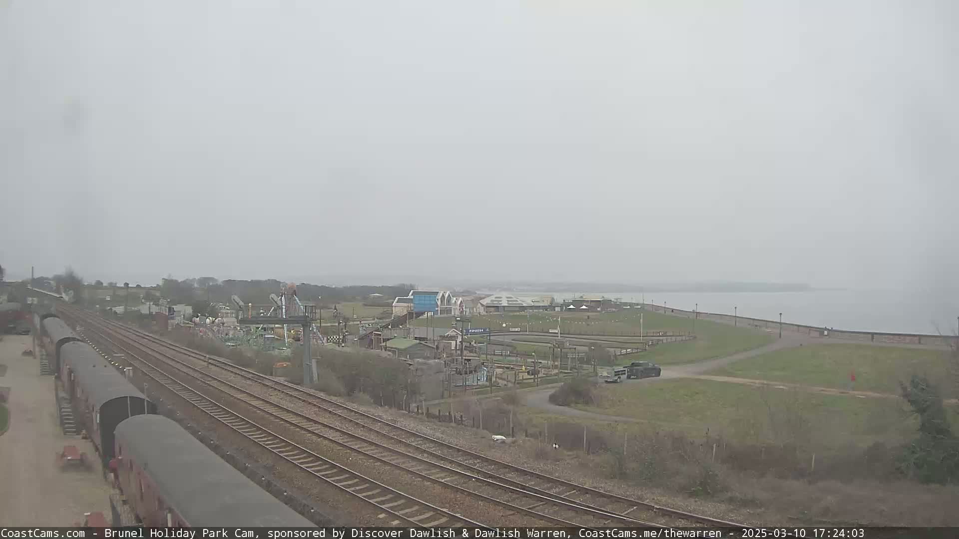 A misty view of a seaside amusement park with train tracks running alongside it, leading to a body of water.