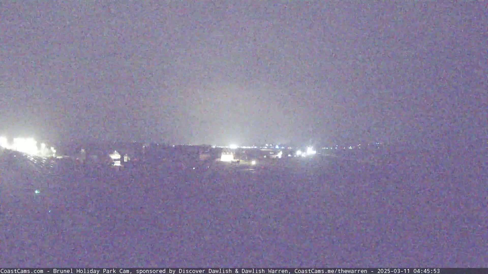 A nighttime long shot of dimly lit buildings and lights in the distance under a dark, overcast sky.