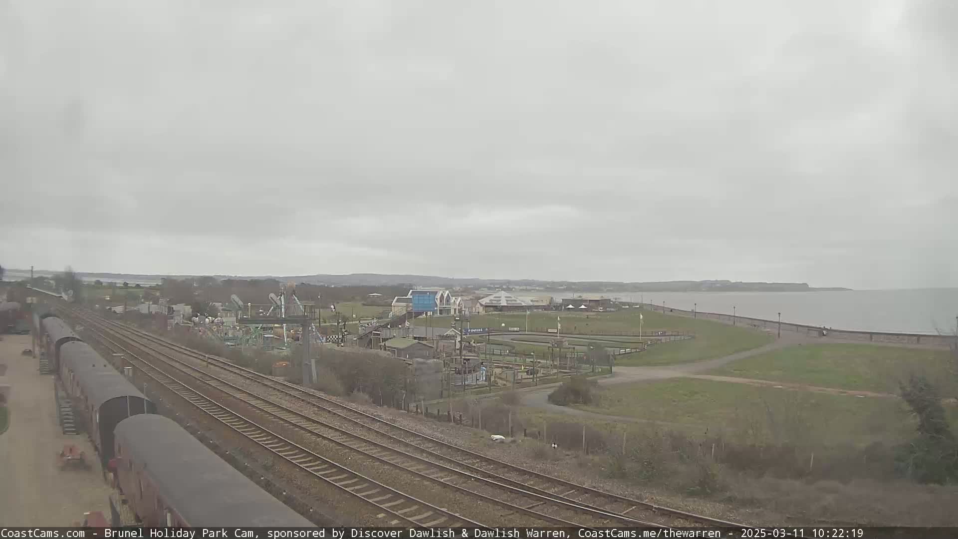 Overcast skies loom over a seaside amusement park and railway lines, with a calm body of water visible in the distance.