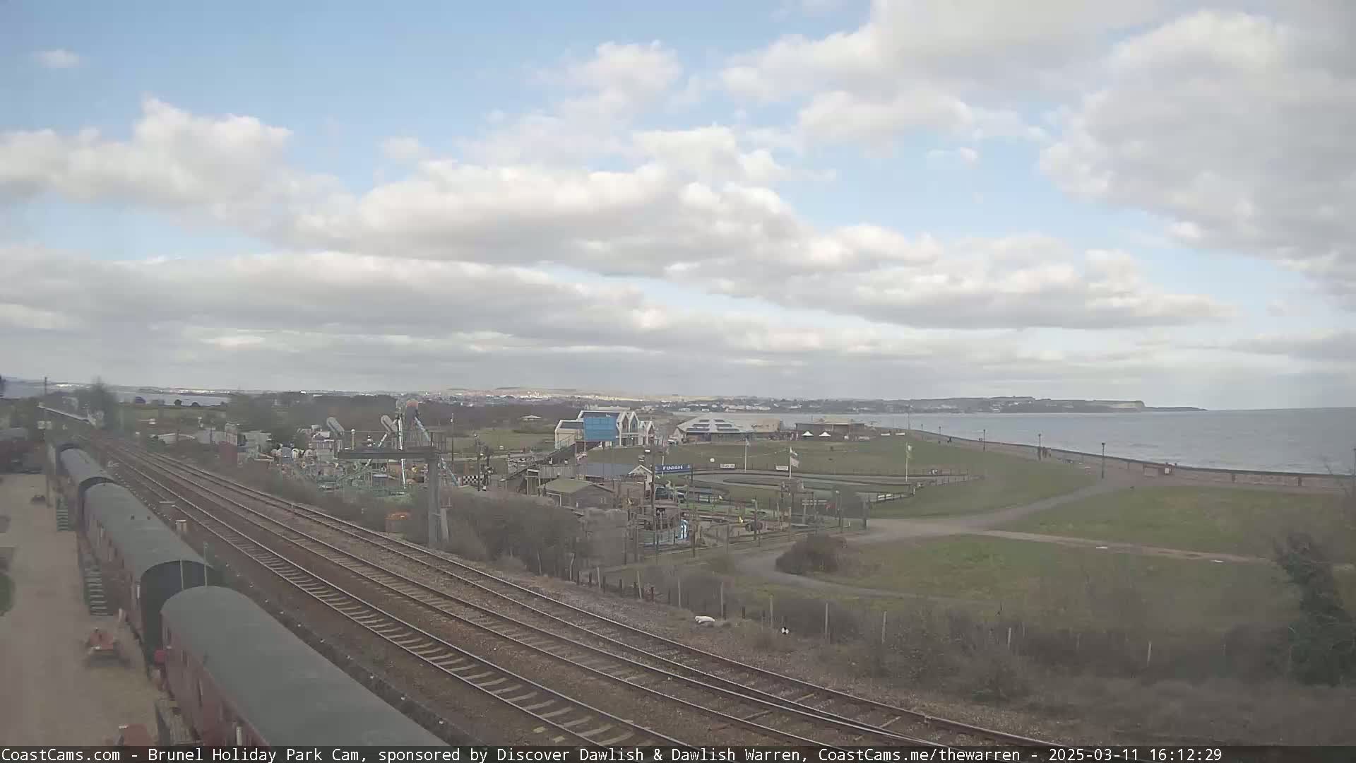 Under a partly cloudy sky, railway tracks run alongside a seaside amusement park toward a calm ocean.