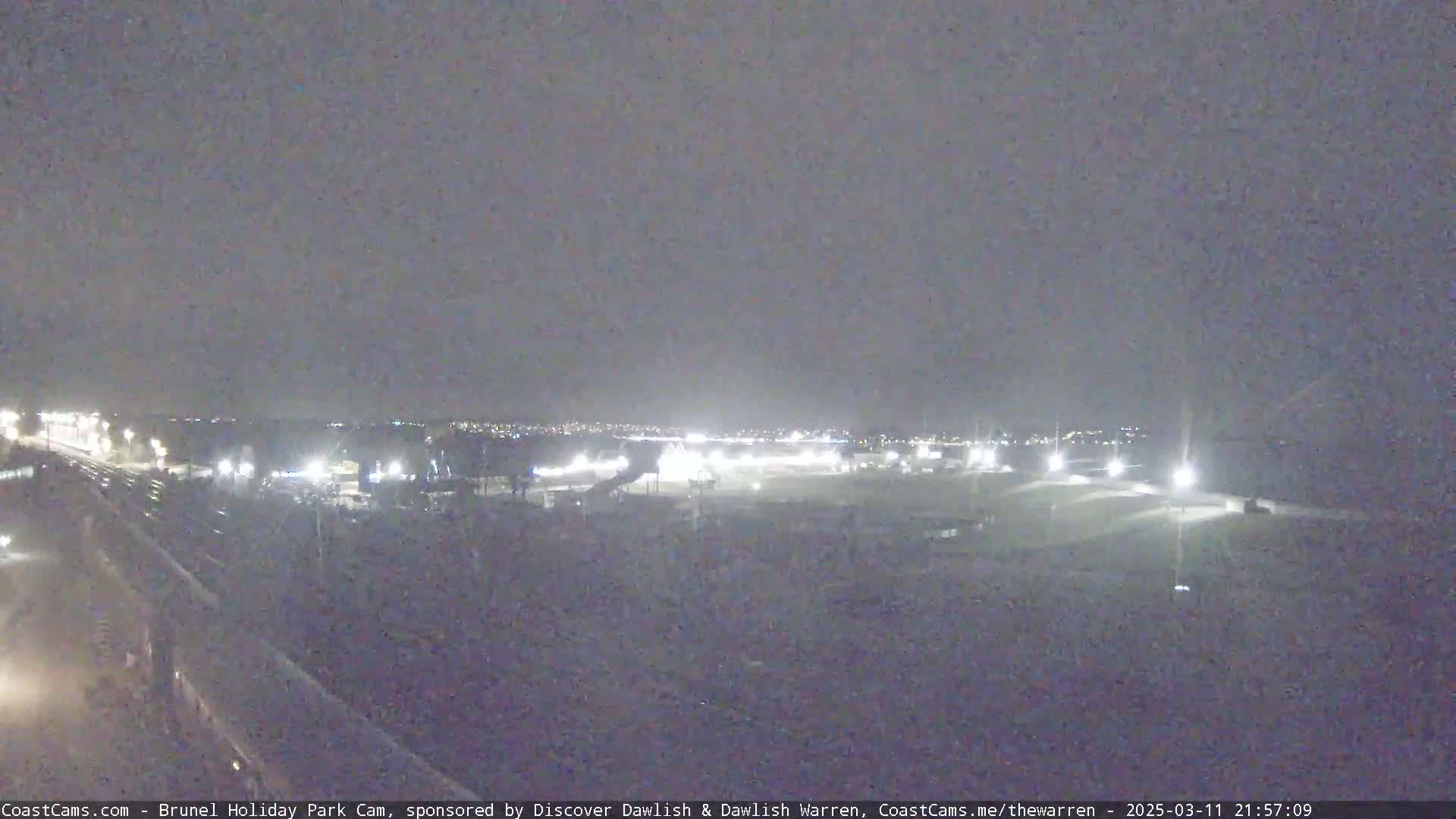 A nighttime, long shot of a mostly empty parking lot and roadway, with distant city lights visible under a cloudy sky.