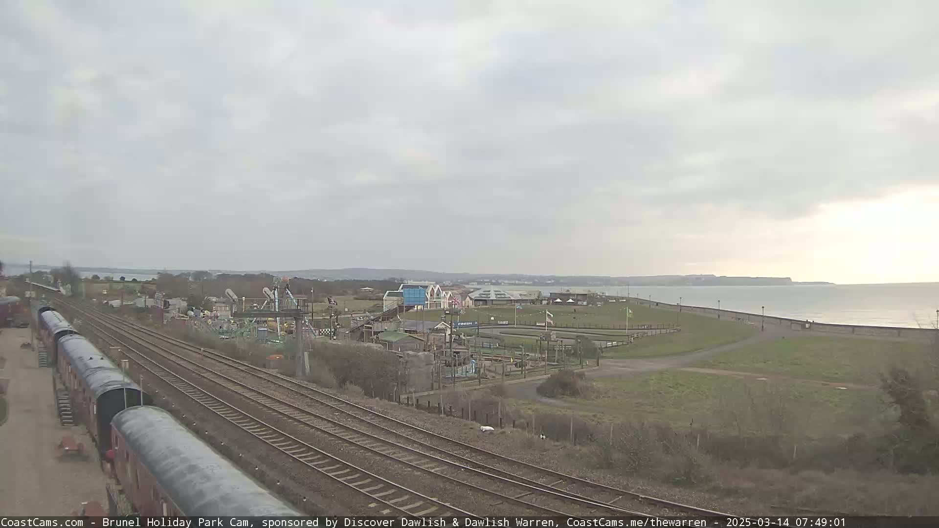 A coastal amusement park and railway lines are visible under an overcast sky, with a calm sea in the background.