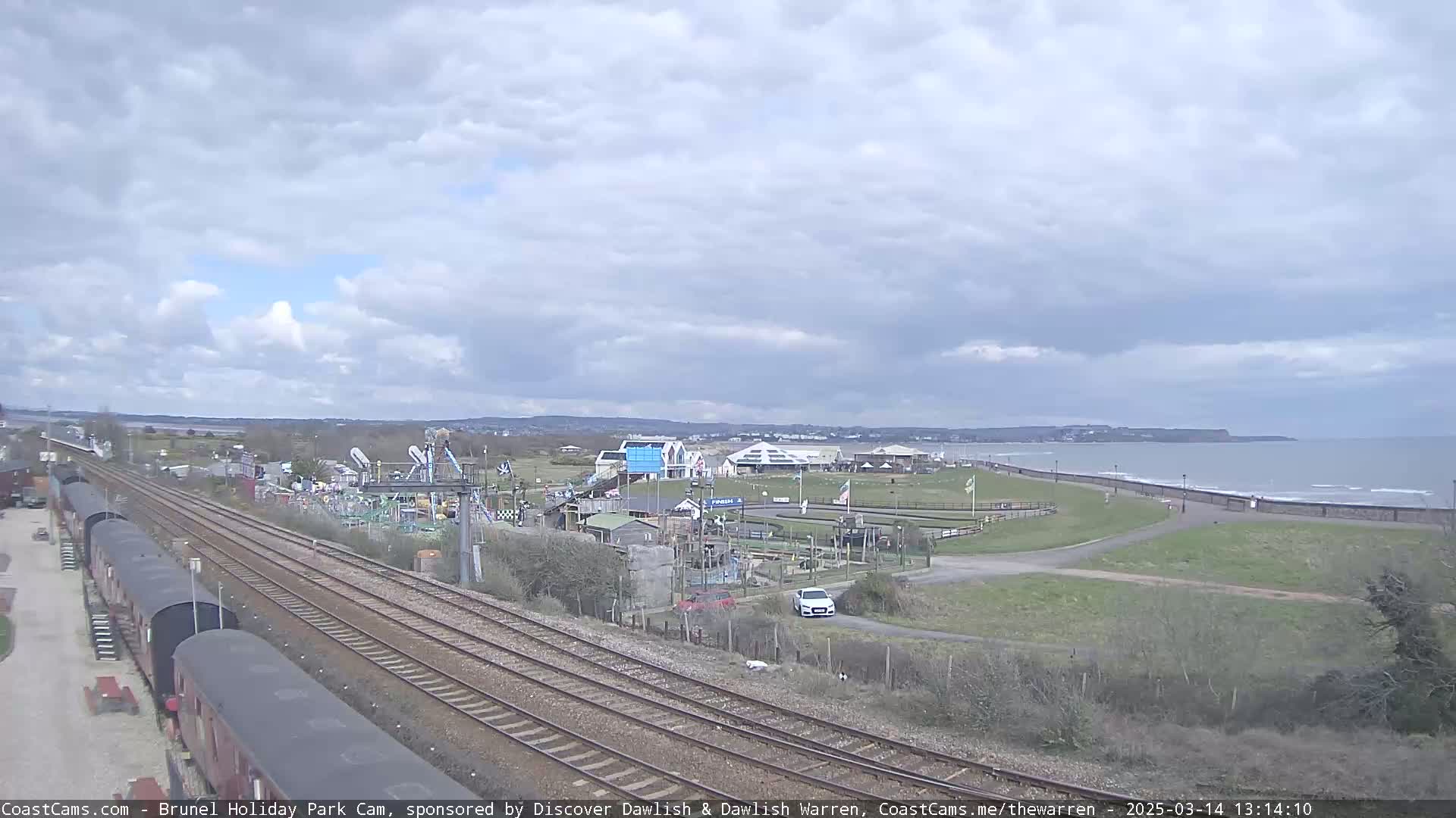 A seaside amusement park with a train track and a cloudy sky is seen overlooking a calm ocean.
