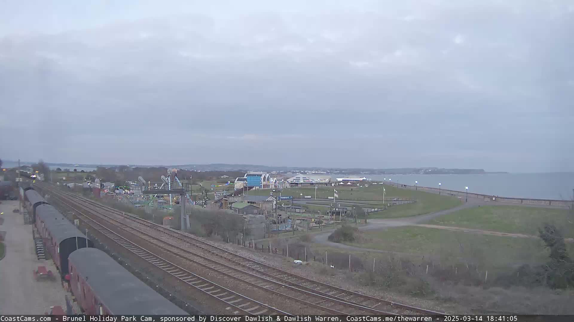 A seaside amusement park is visible next to railroad tracks under an overcast sky.