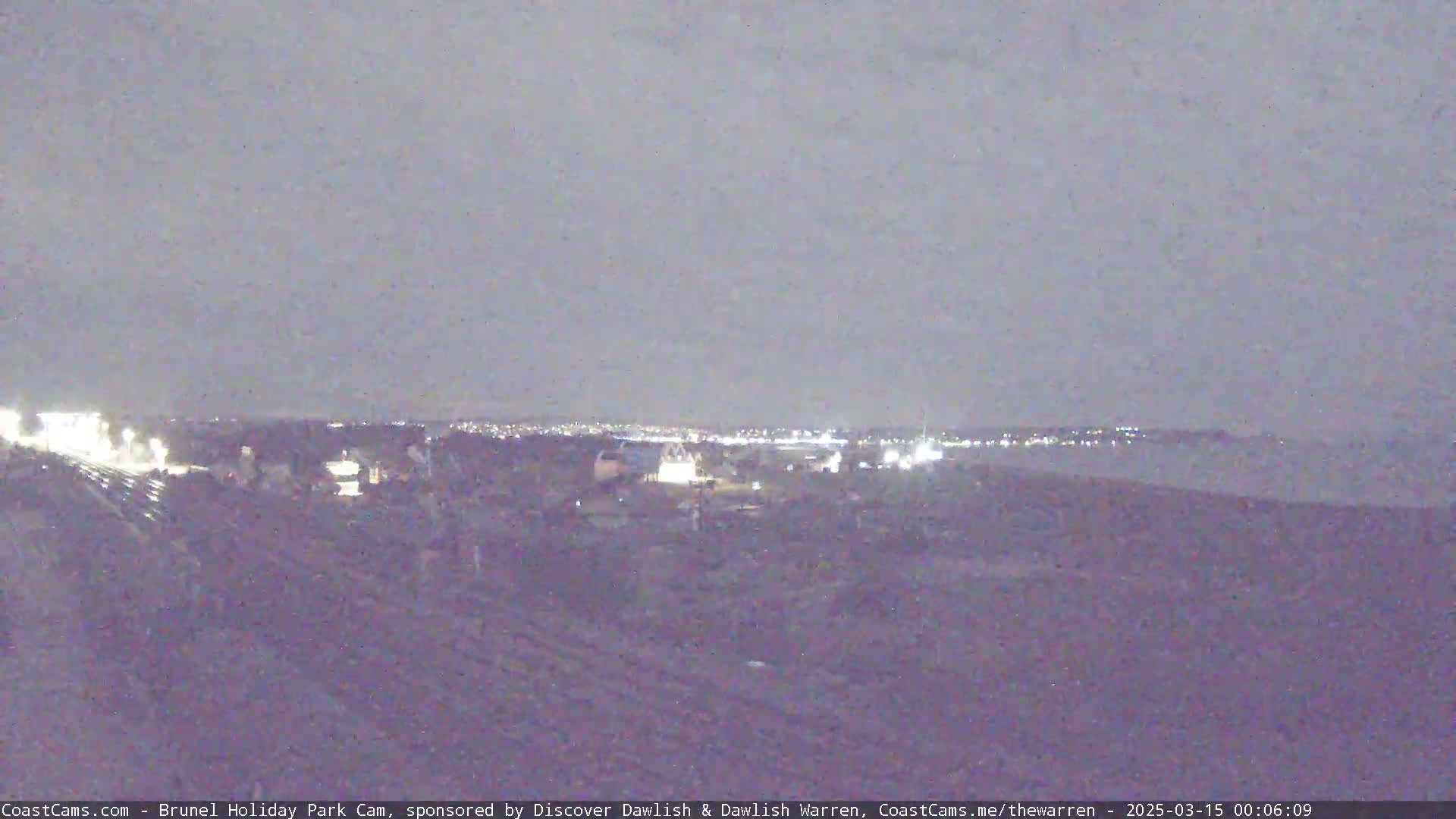 A nighttime long shot shows a dimly lit landscape with a distant town's lights visible on the horizon under a dark, overcast sky.