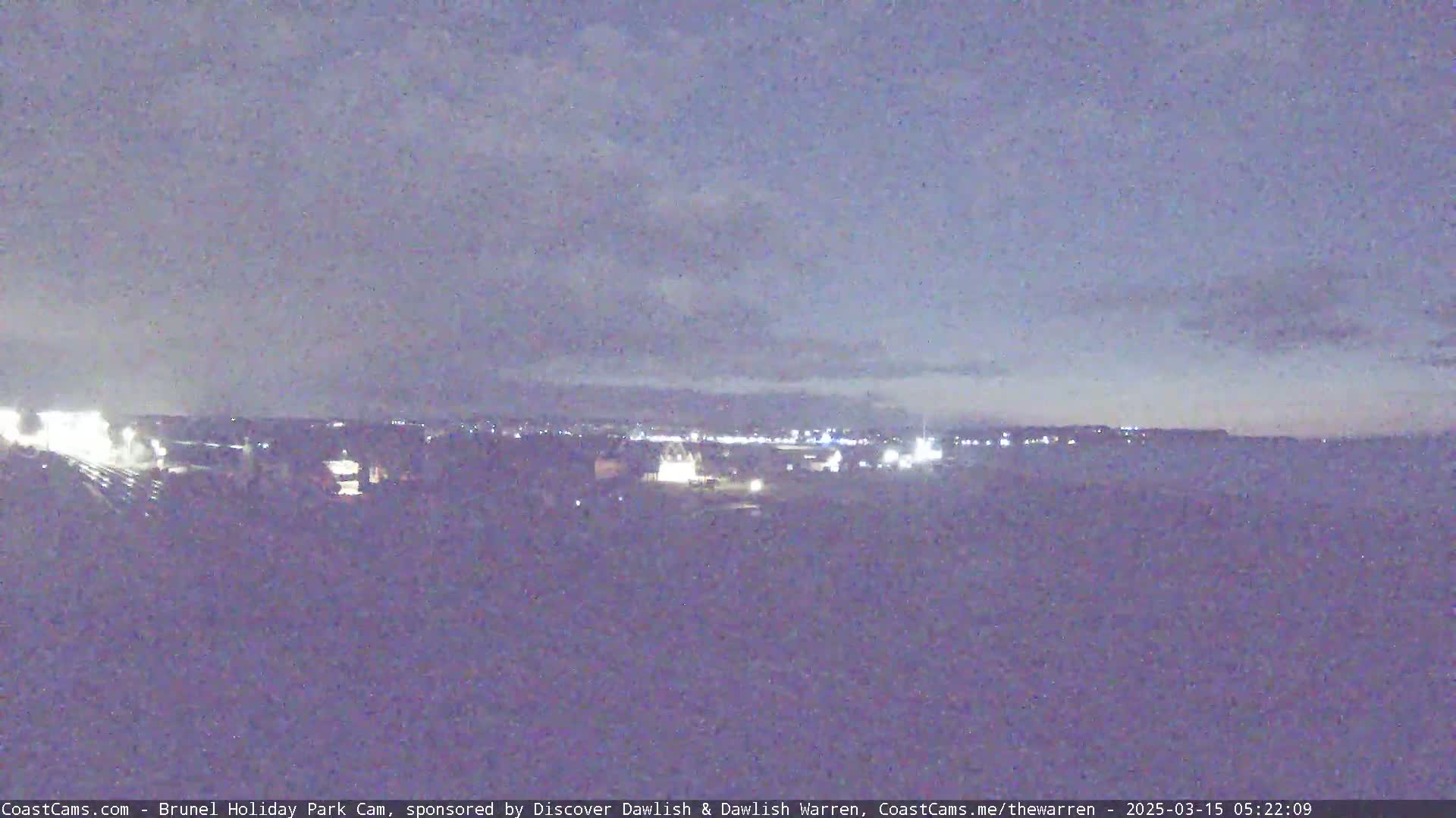 A nighttime long shot shows a distant town with lights under a dusky, cloudy sky.