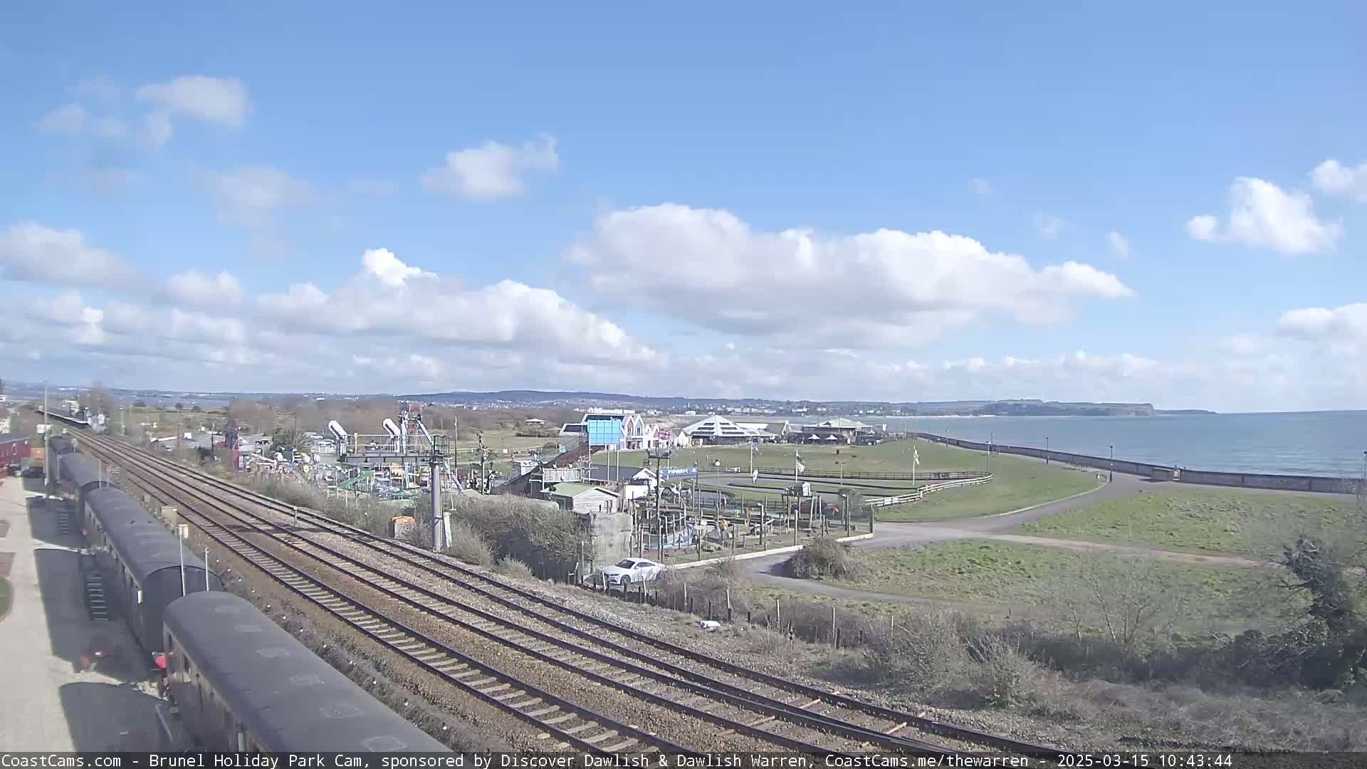 A train track runs alongside a seaside amusement park on a partly cloudy day, with a calm ocean visible in the distance.