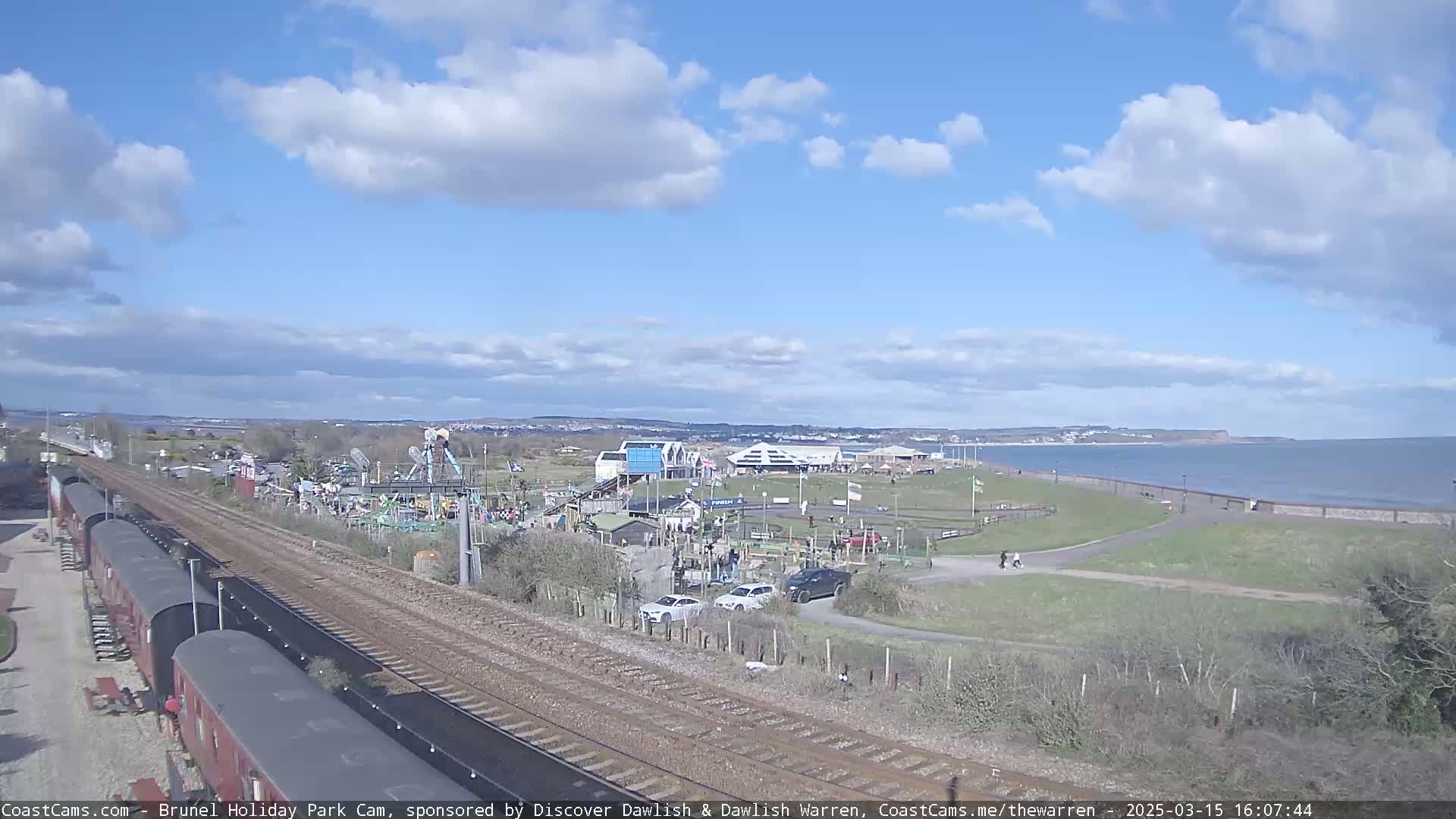 A seaside amusement park with a train track running alongside, under a partly sunny sky with scattered clouds.