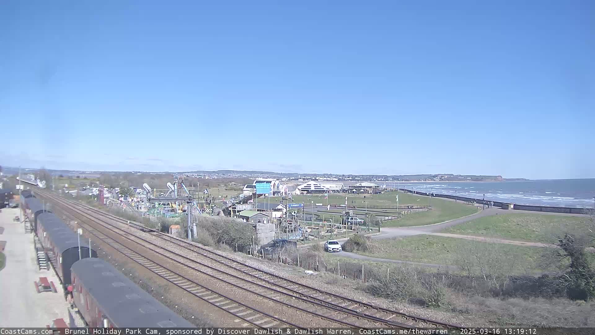A seaside amusement park with a go-kart track and railway lines is visible on a sunny day with a calm ocean in the background.