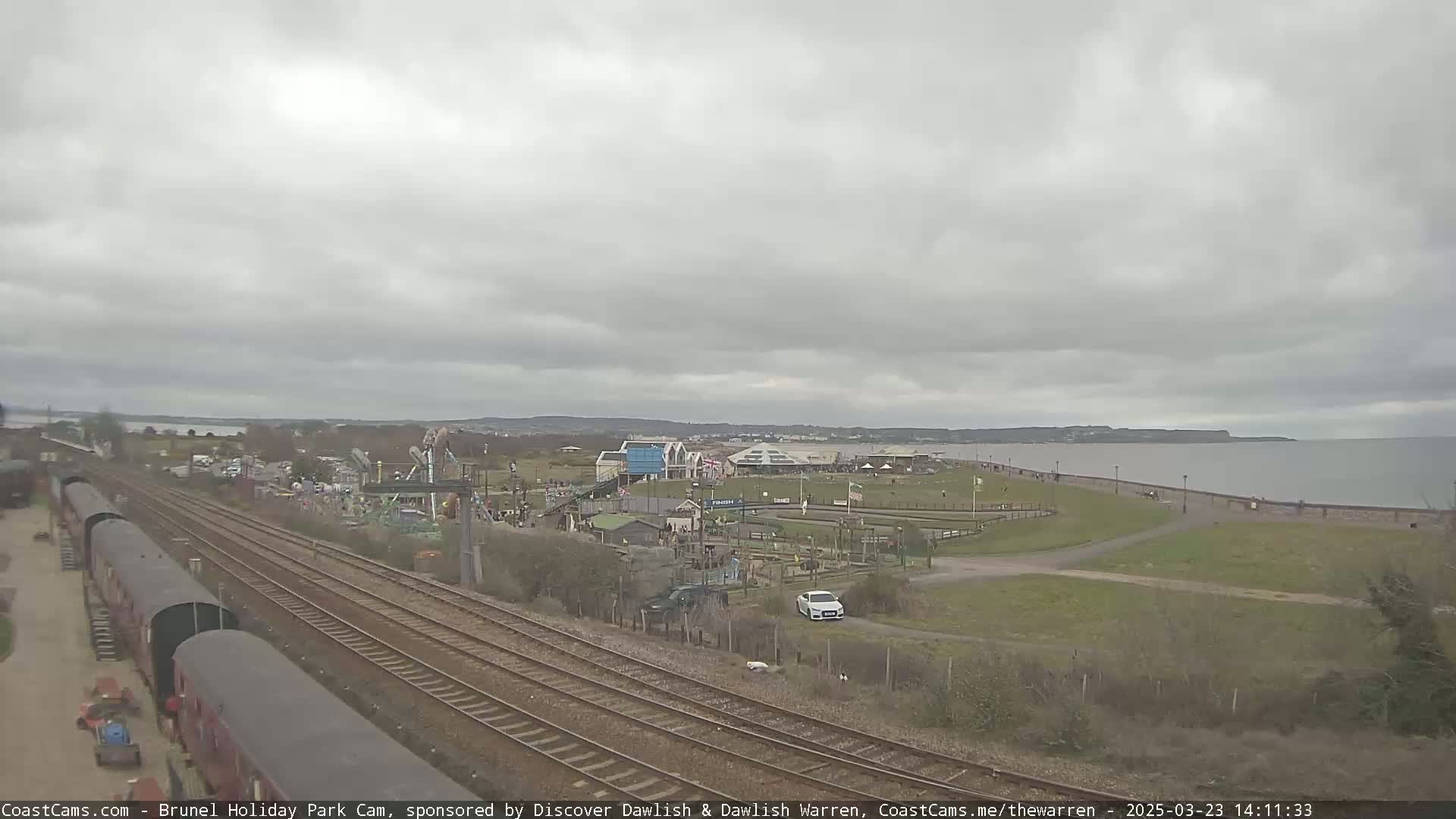 A coastal amusement park with railway tracks nearby is viewed under an overcast sky, next to a calm body of water.