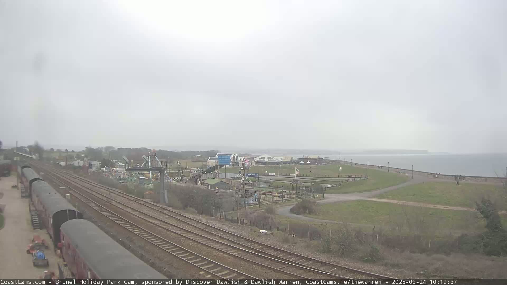 A dreary, overcast day shows a coastal area with railway tracks, a holiday park featuring amusement park rides and go-karts, and a calm body of water in the distance.