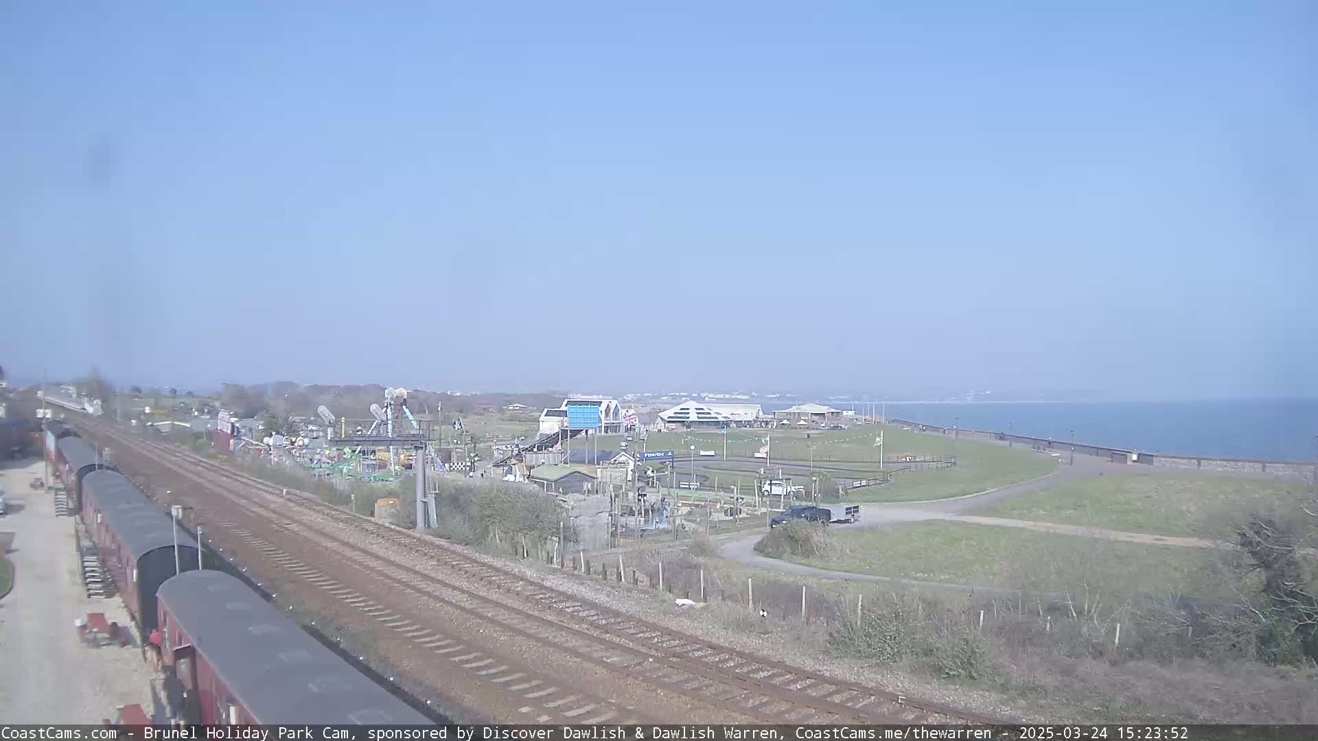 A sunny day overlooks a seaside amusement park with train tracks and several parked railway carriages visible in the foreground.