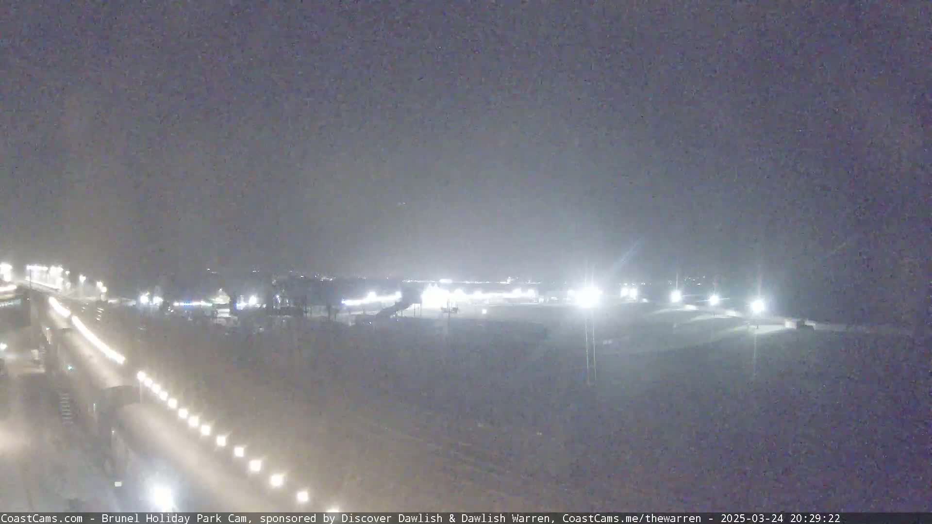 A nighttime, dimly lit view of a roadway and a large, mostly dark parking lot with some lights visible.