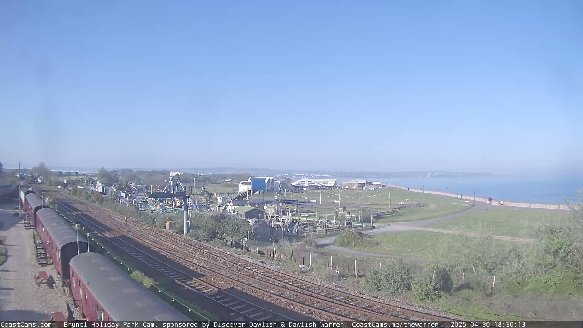 A sunny seaside scene shows train tracks alongside a coastal amusement park and a calm ocean.