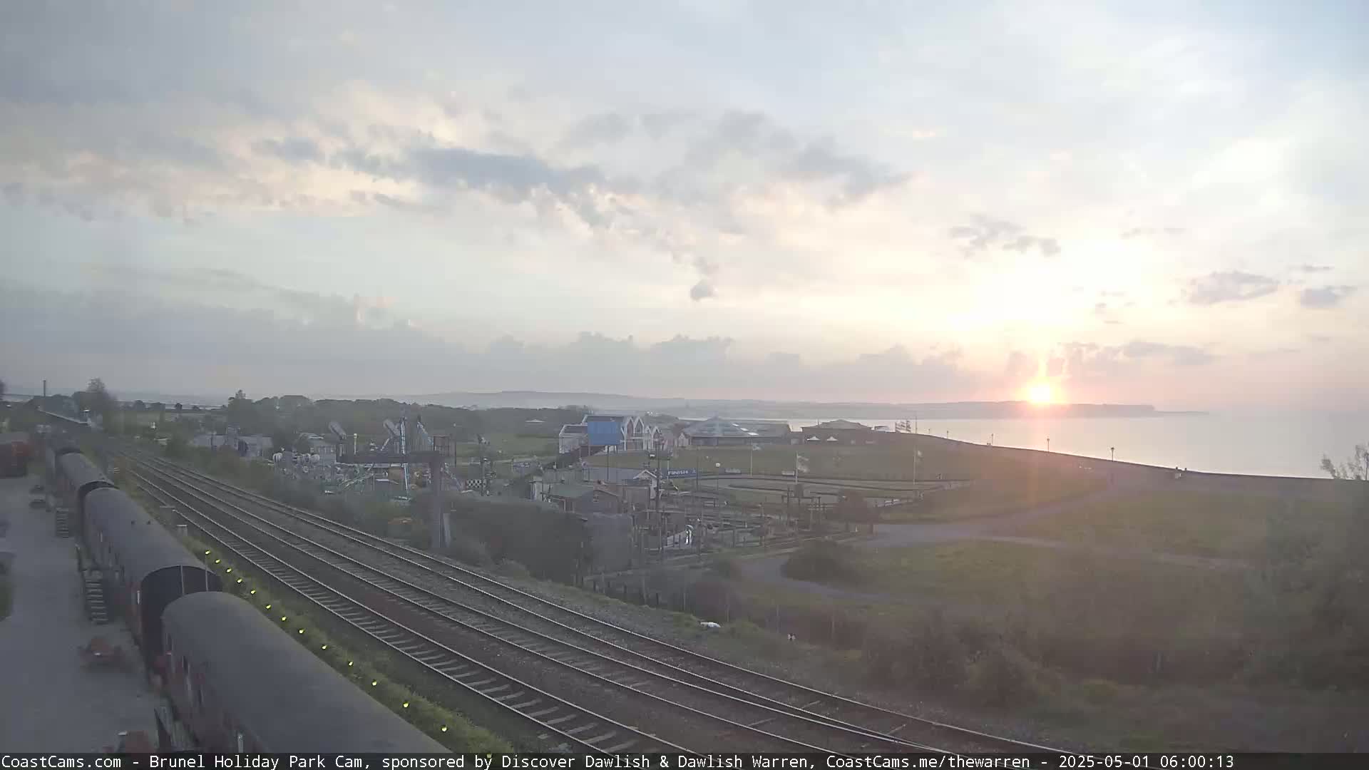 A sunrise over a calm ocean is visible from a vantage point overlooking railway tracks and a seaside town.