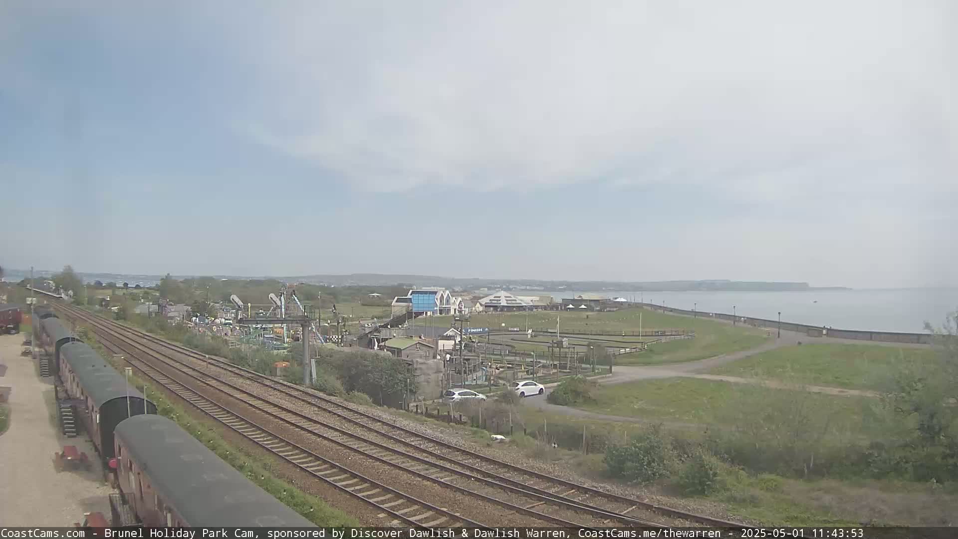 A seaside amusement park with train tracks and railway cars is visible under a partly cloudy sky, next to a calm body of water.