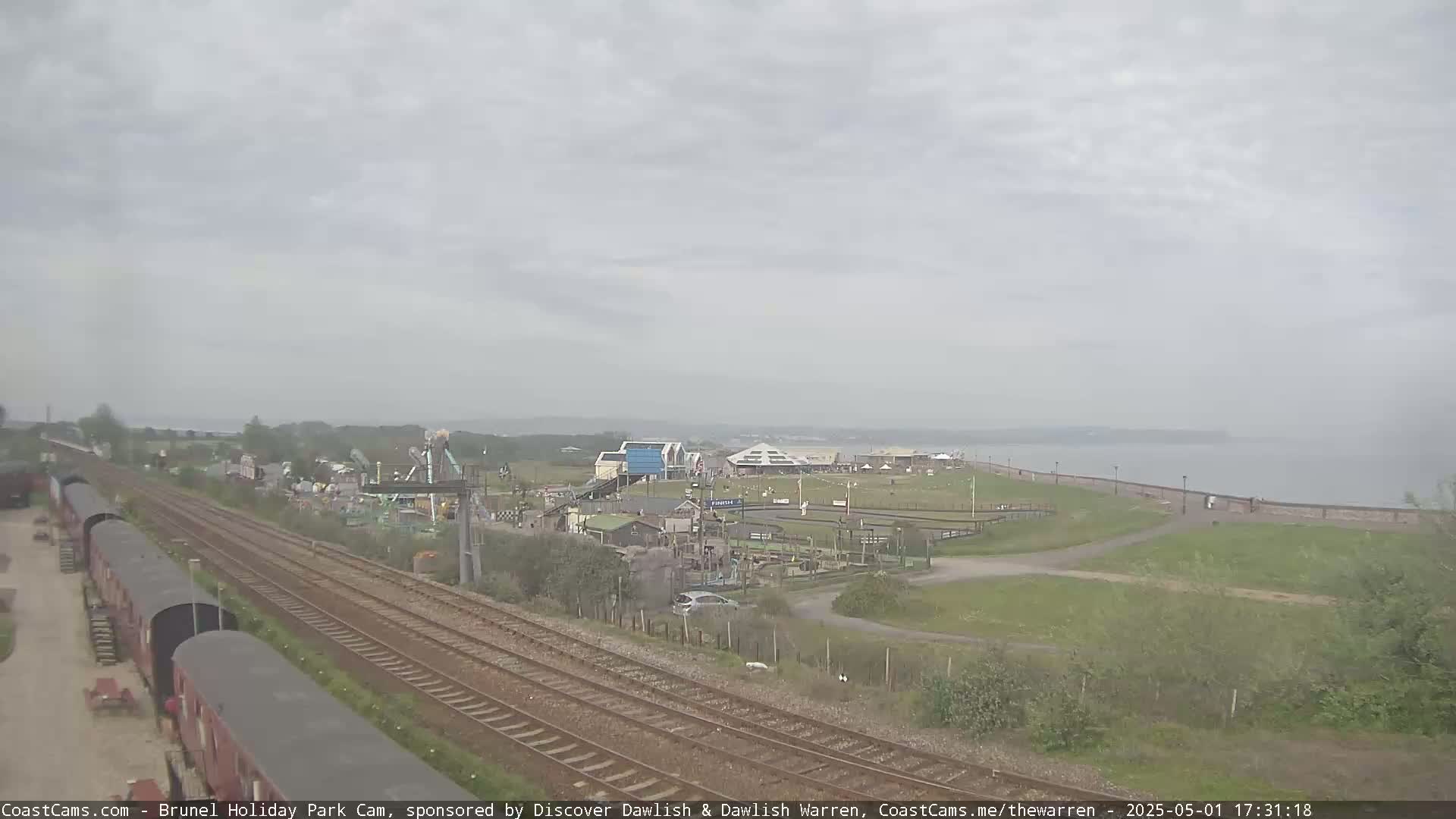 A seaside amusement park is visible from a high vantage point, with overcast skies and railway tracks in the foreground.