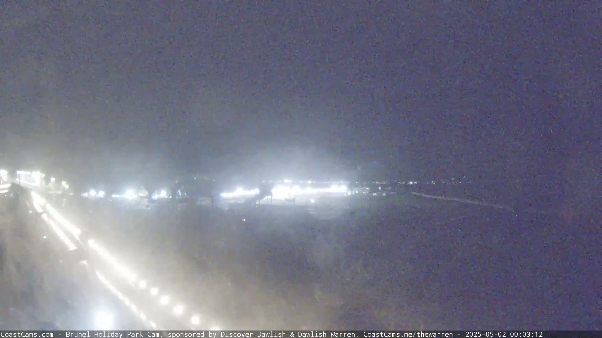 A nighttime, hazy view of a roadway with headlights visible, and brightly lit structures in the distance.