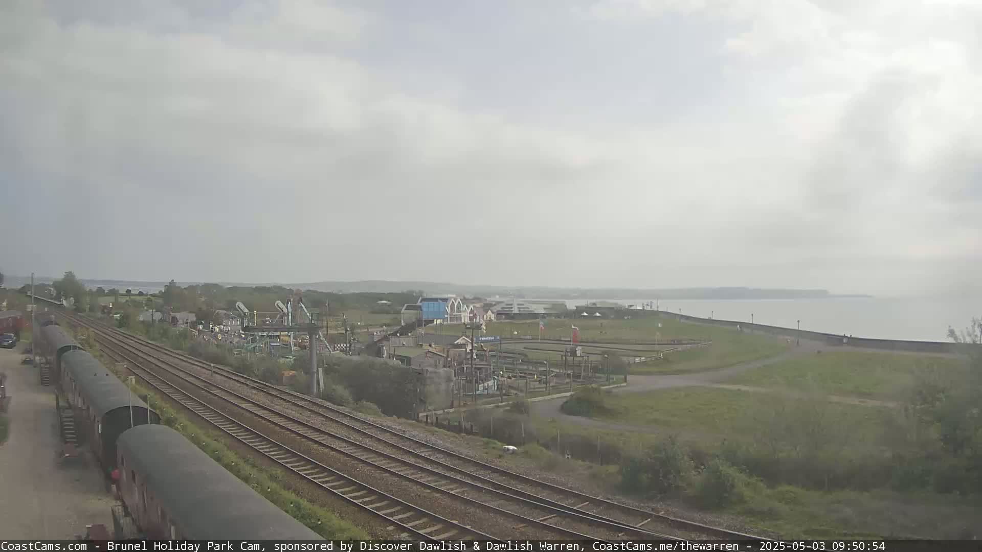 A coastal view on an overcast day shows railway tracks leading to a park with amusement-style buildings and a calm sea in the background.