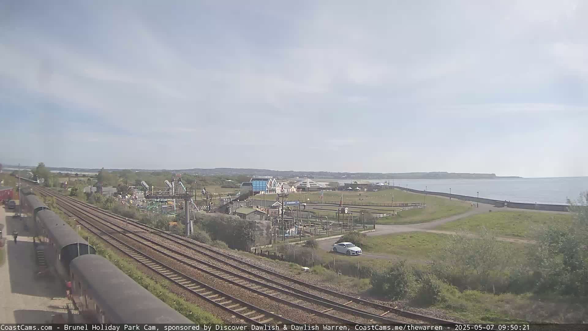 A seaside amusement park with a go-kart track and railway lines is visible, under a mostly sunny sky, near a calm body of water.