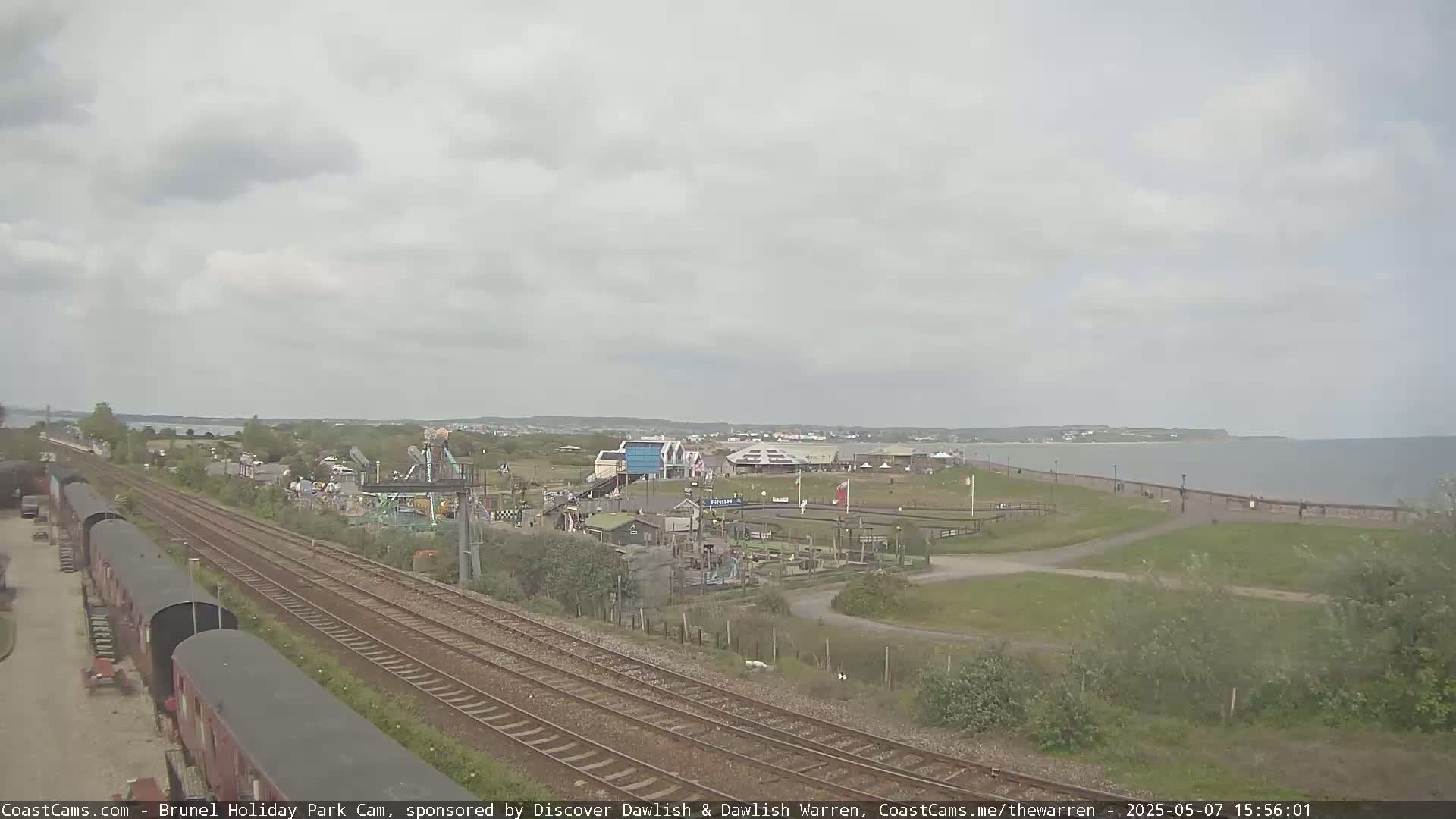 An overcast day at a seaside amusement park, with train tracks running alongside it and a calm body of water visible in the distance.