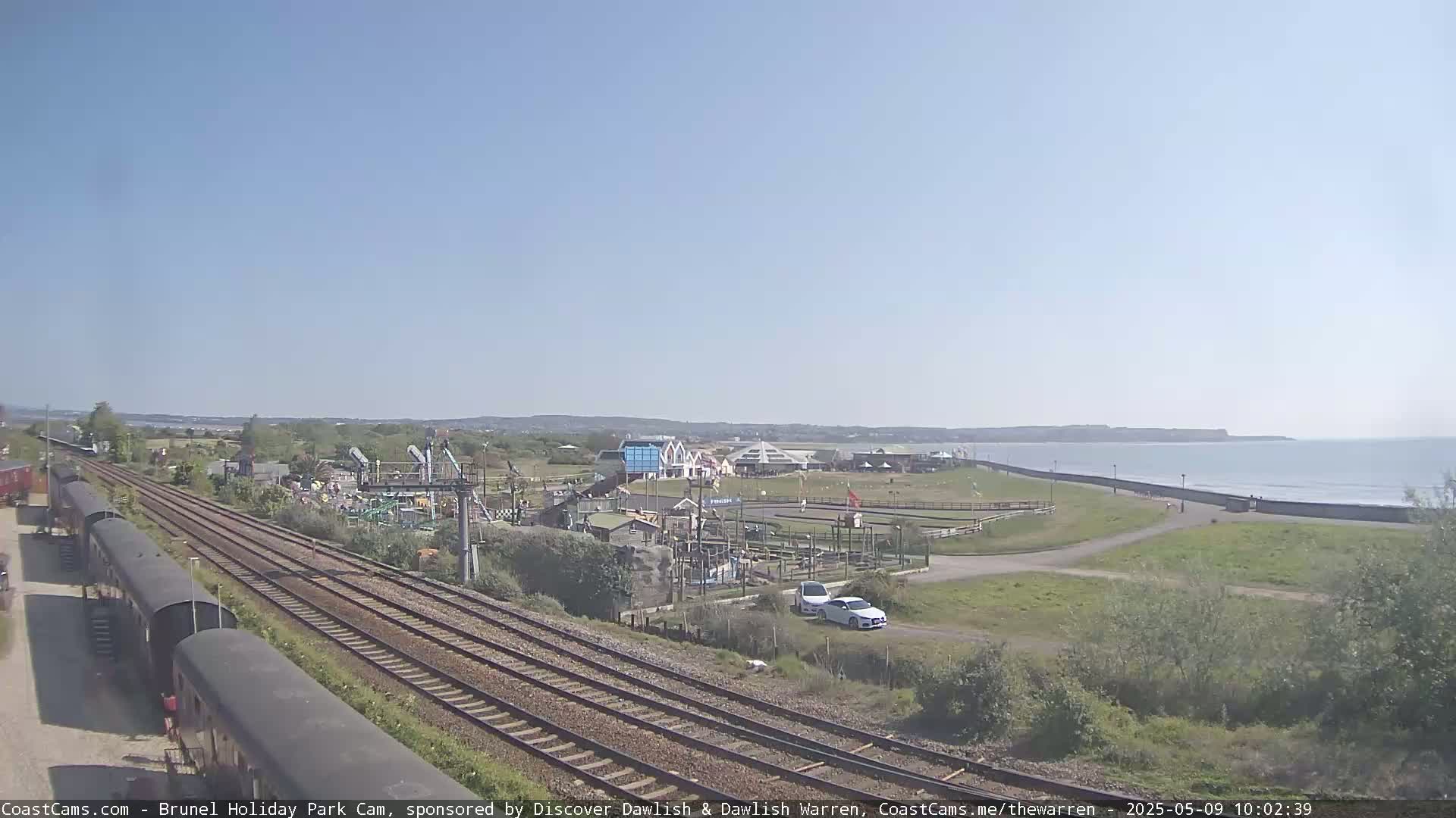 Under a clear blue sky, a seaside amusement park is visible beyond railroad tracks and several parked railway cars.