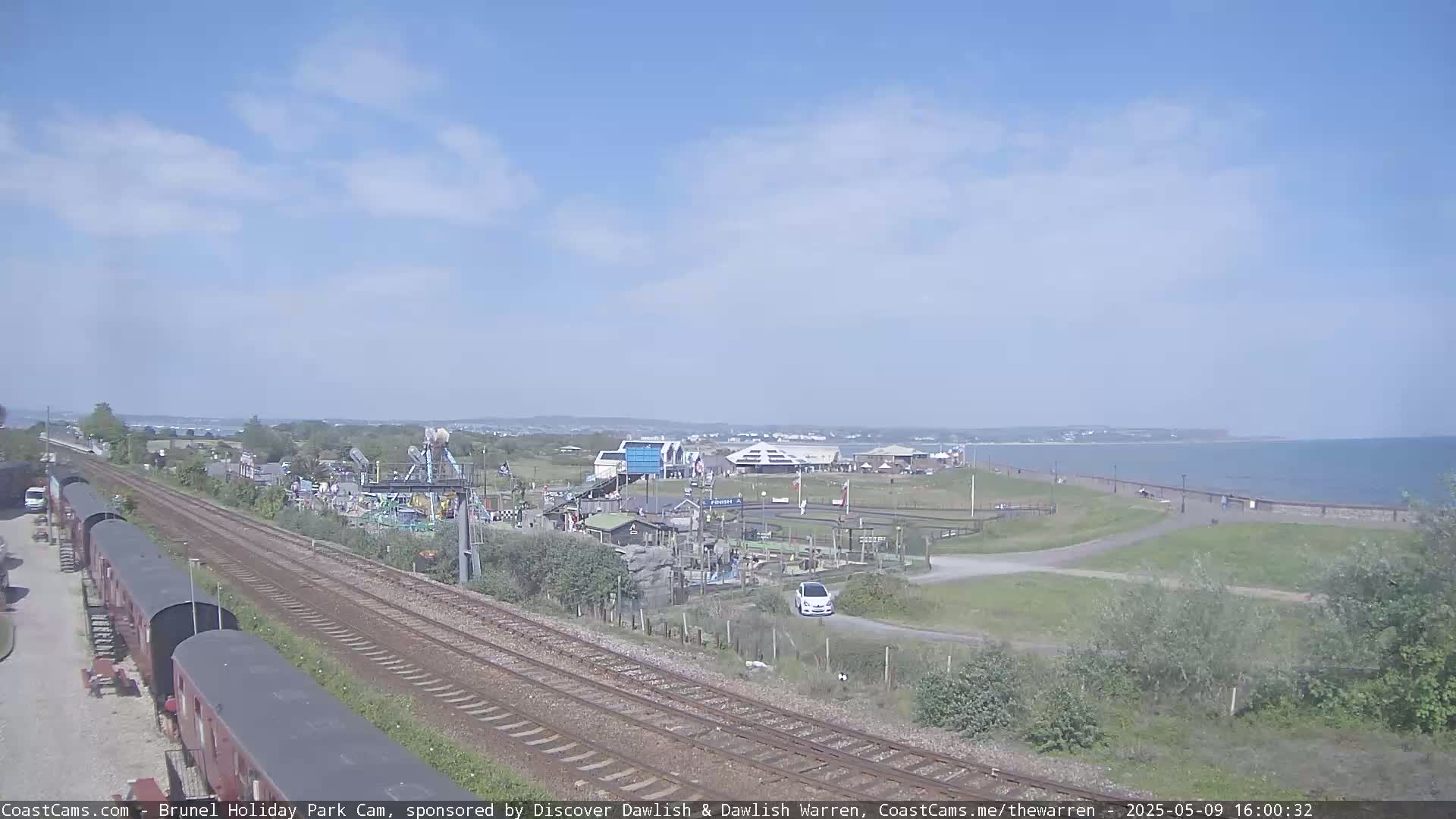 A seaside amusement park with a small car and train cars is visible near a calm ocean under a mostly sunny sky.