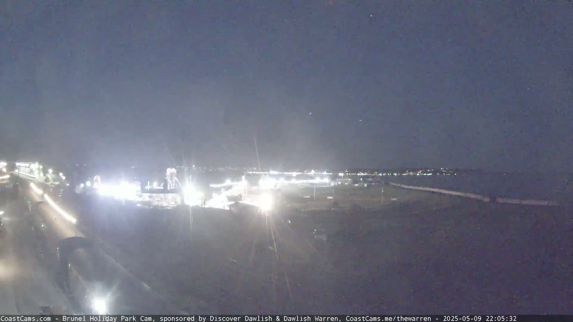 A nighttime long shot of a coastal area shows a road with car lights, a brightly lit amusement park area, and a dark body of water with faintly lit buildings in the distance under a clear, dark sky.